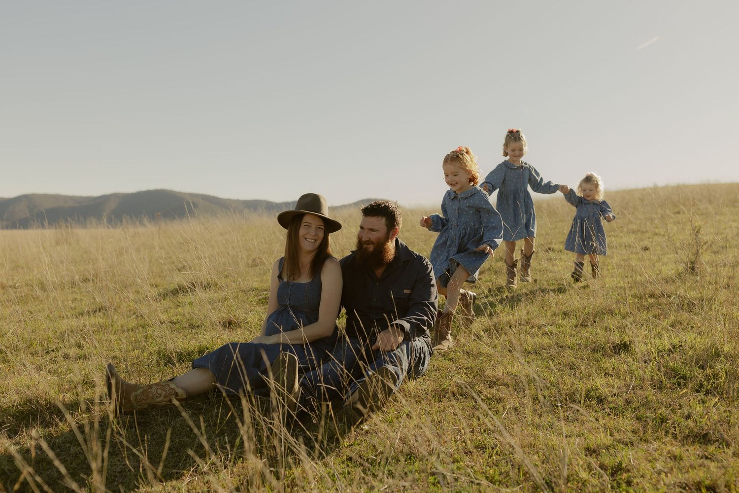 Family sitting and playing in a sunny green meadow with rolling hills, parents relaxing while children dance joyfully.