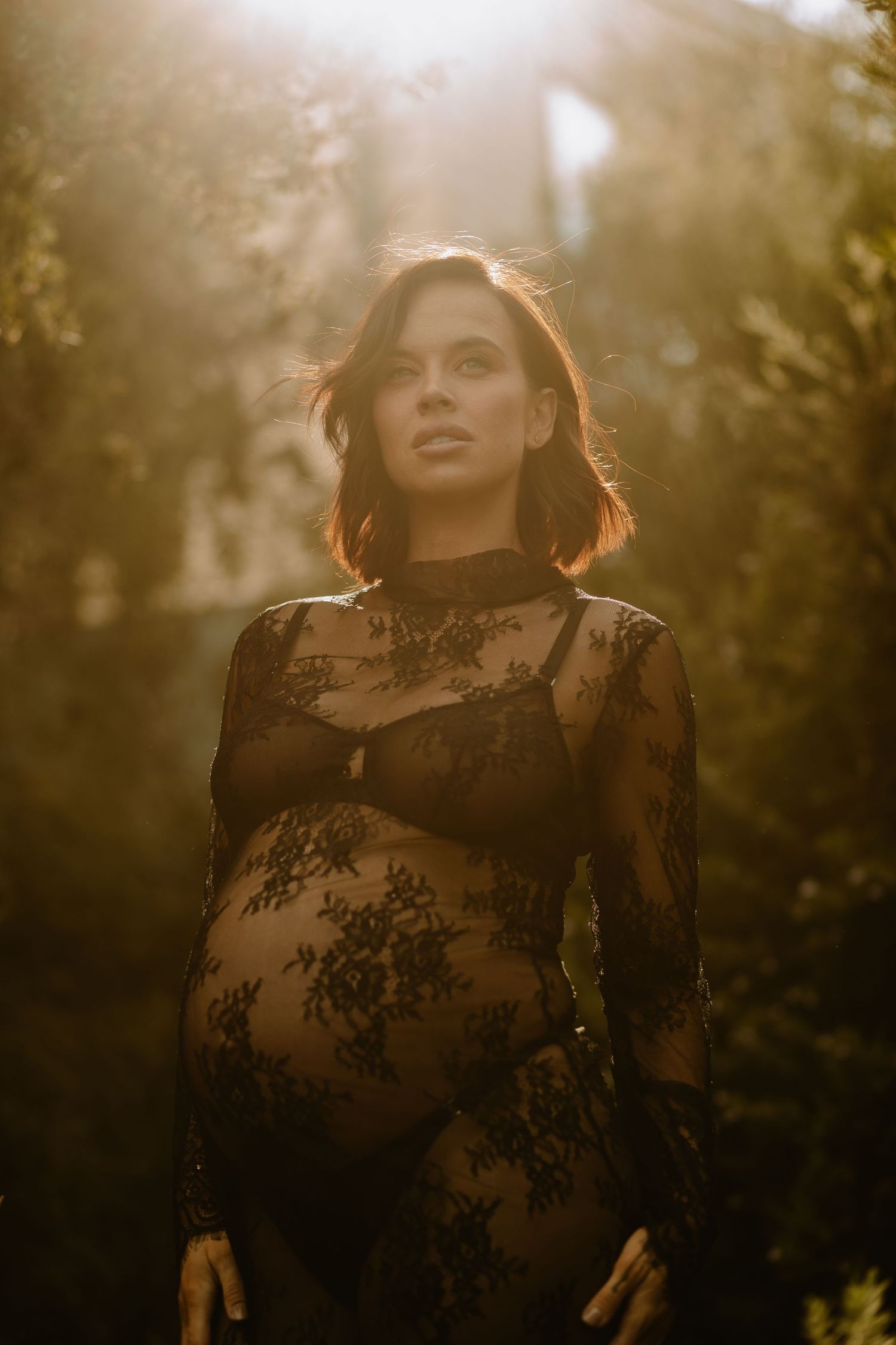 An artistic maternity portrait in a sheer lace dress with golden sunlight filtering through trees.