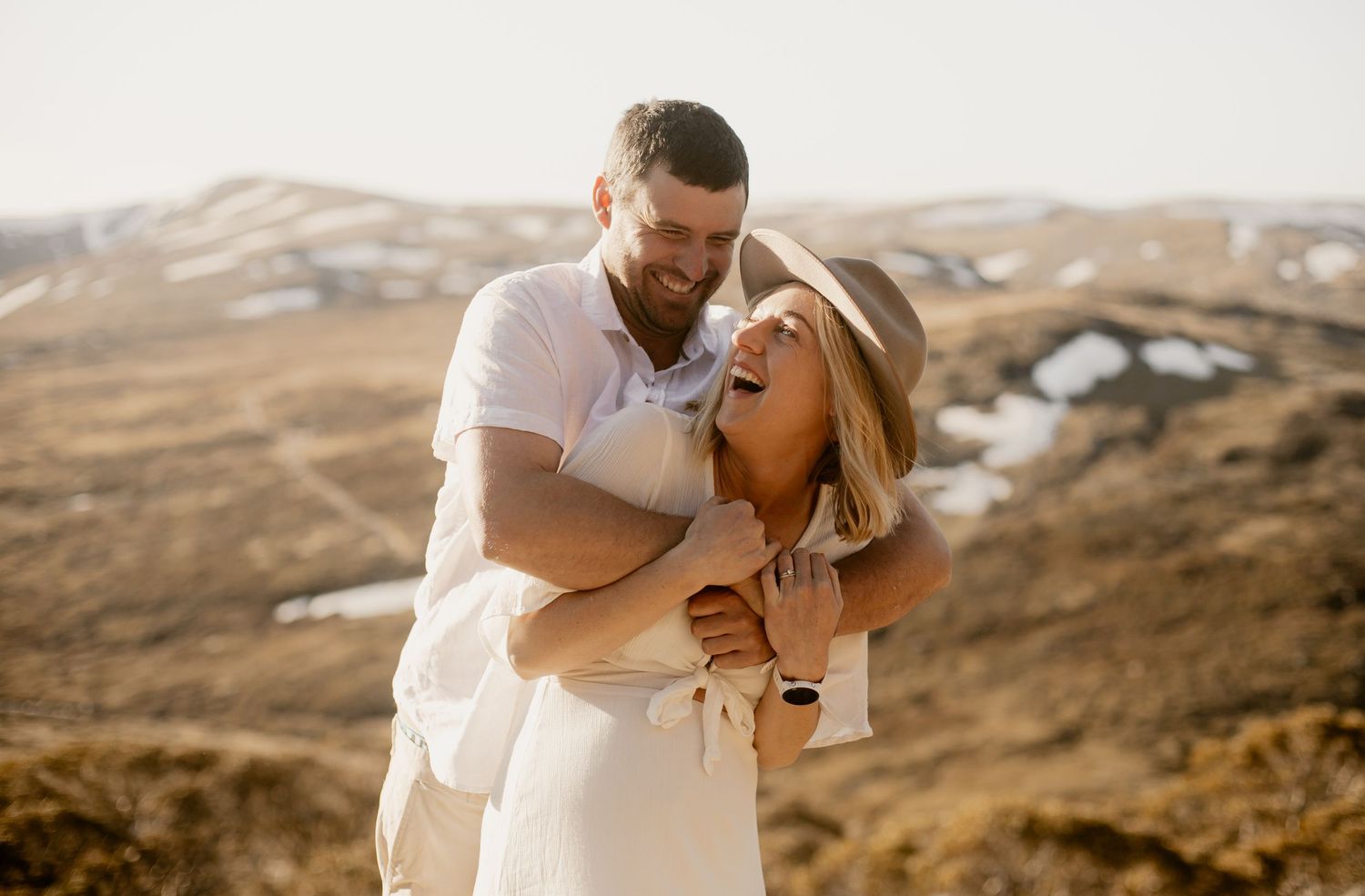 A joyful couple embraces and laughs together against a backdrop of rolling desert hills during golden hour.
