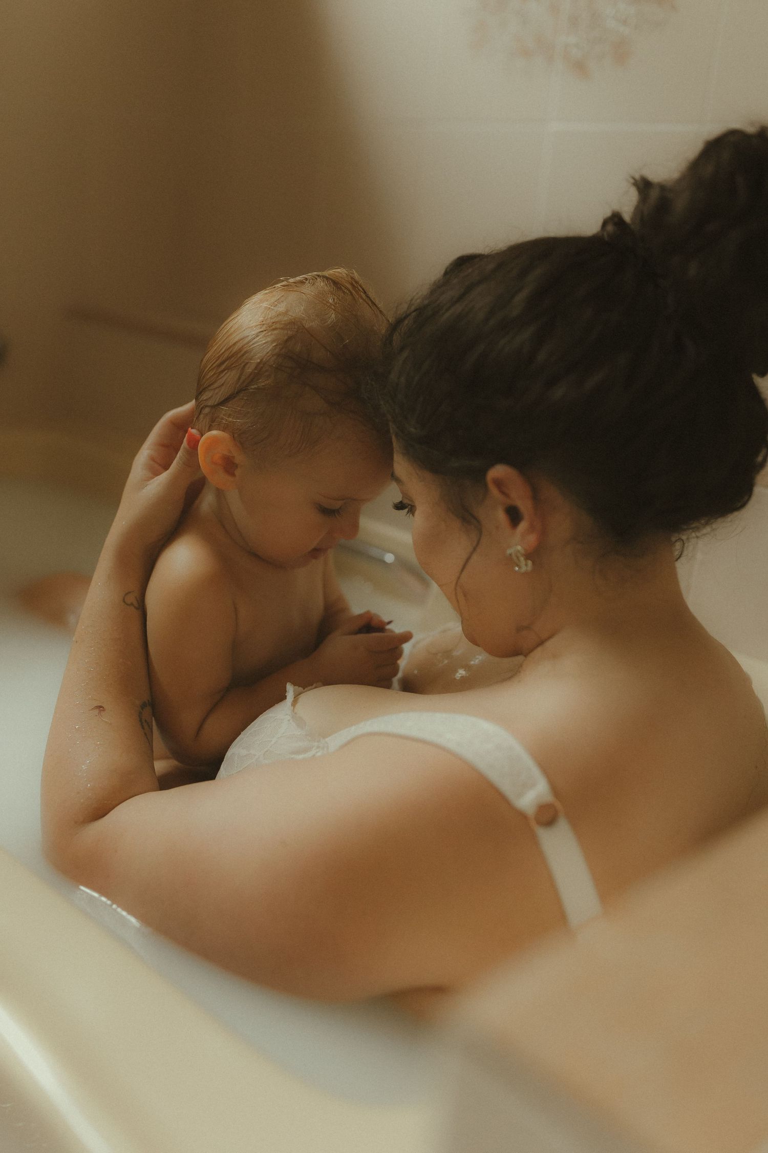 Intimate bathtub portrait of a tender bonding moment between parent and baby in soft warm lighting.