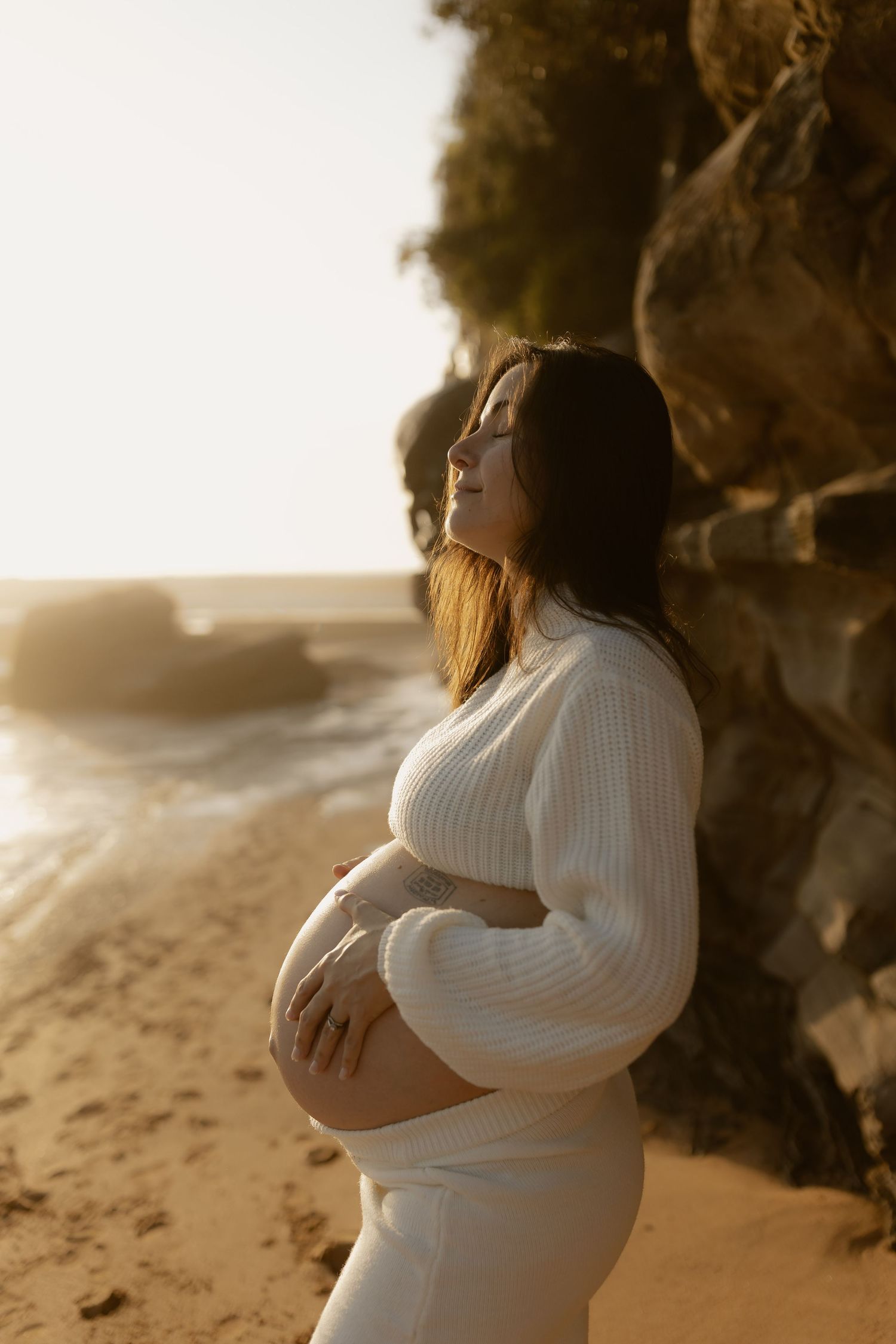 Silhouetted figure in white sweater poses on beach at sunset while cradling pregnant belly.