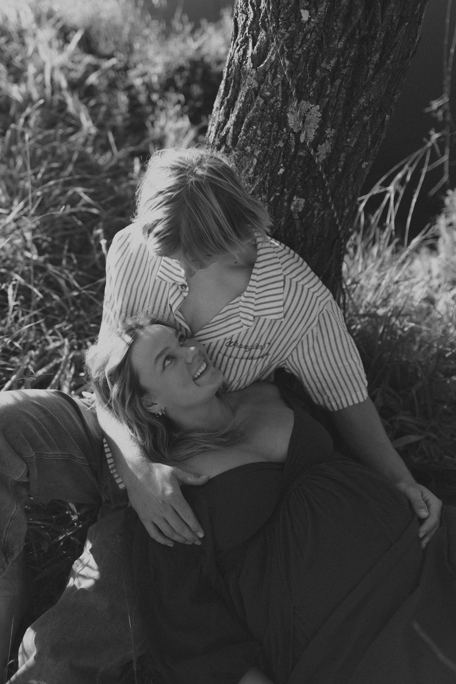 Intimate black and white portrait of two people resting against a tree trunk.
