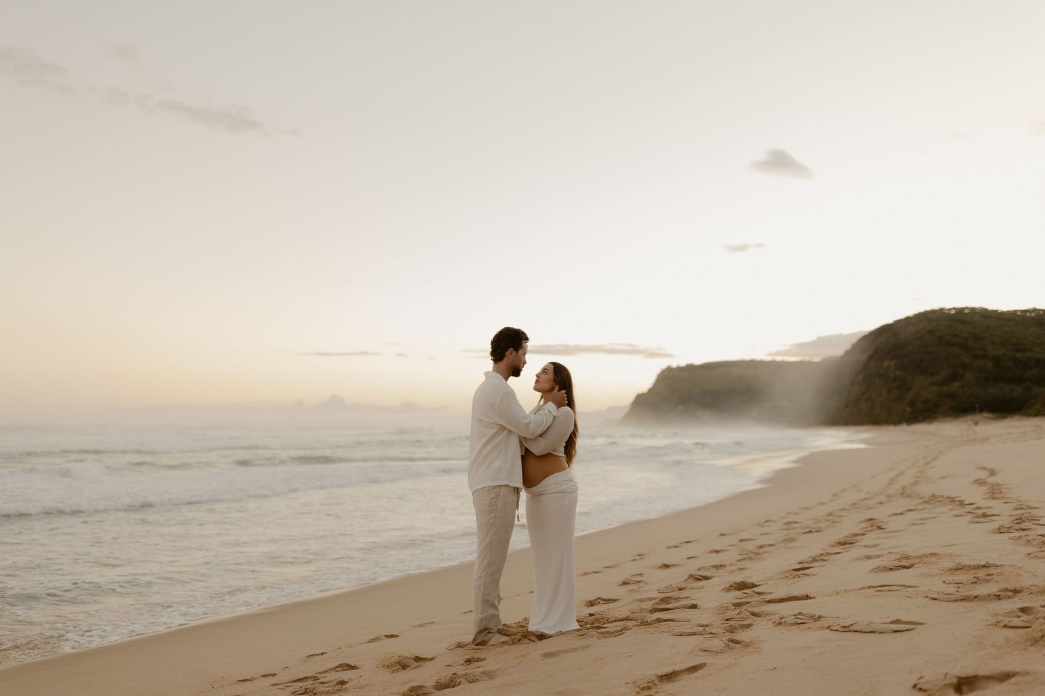 A couple in white clothing walks along a scenic beach at sunset with mountains in the background.