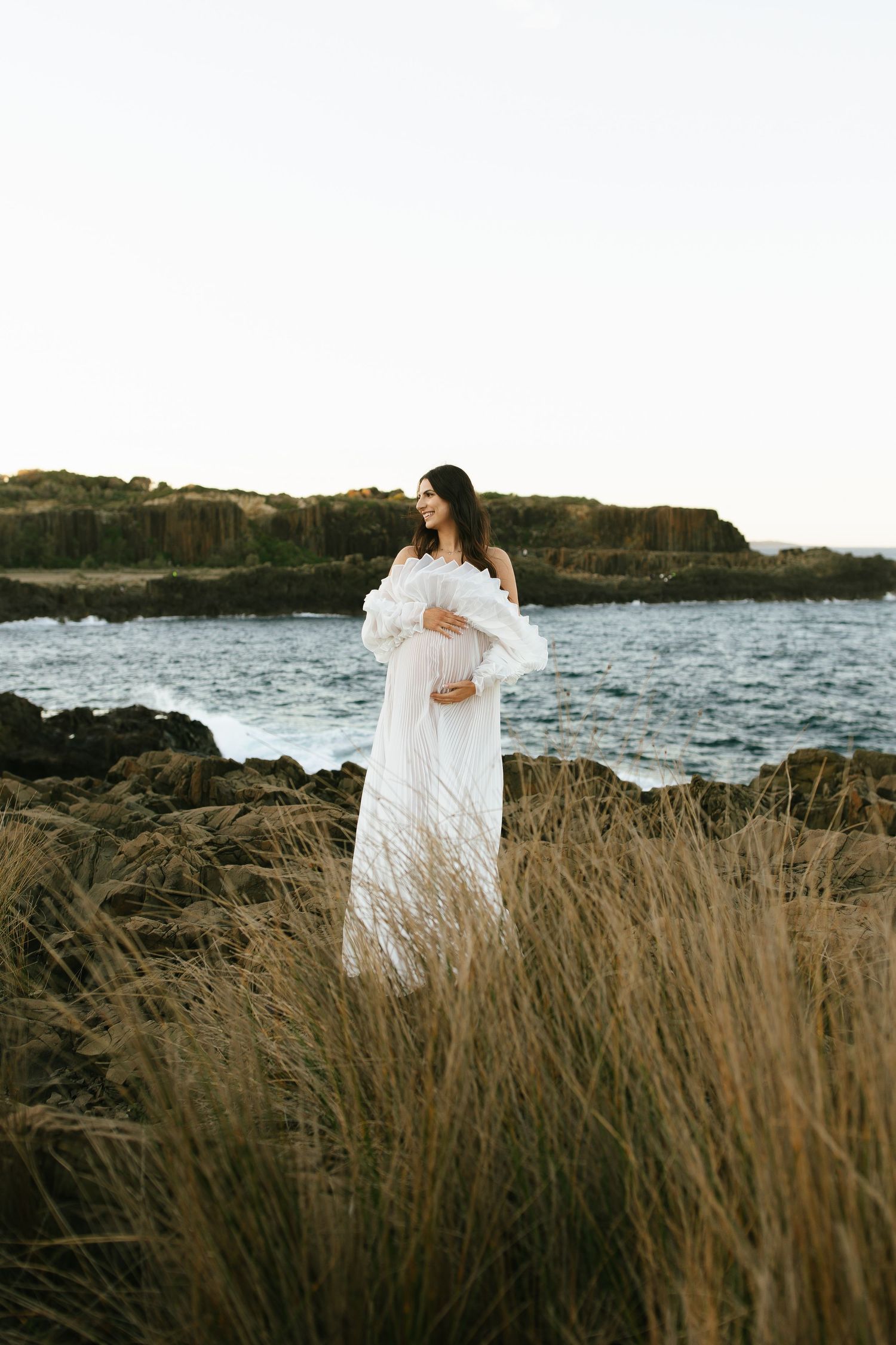 A figure in flowing white dress stands among tall coastal grasses with ocean and rocky cliffs in background.