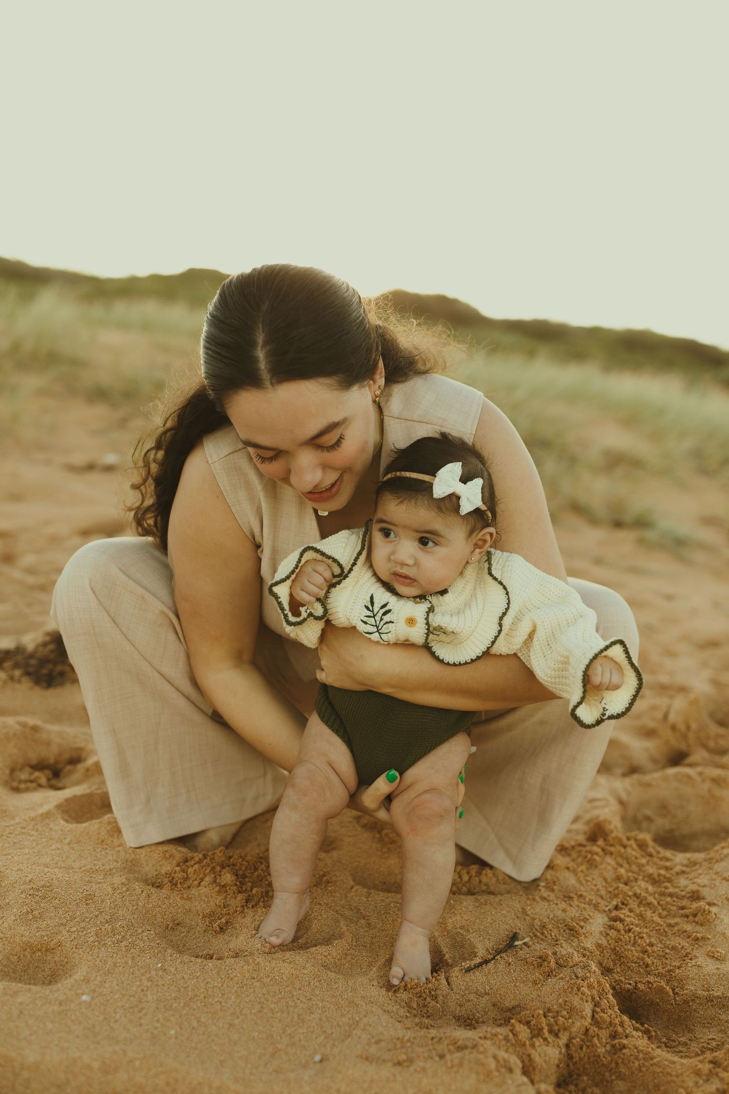 A mother and baby share a tender moment together on a sandy beach in warm sunlit conditions.