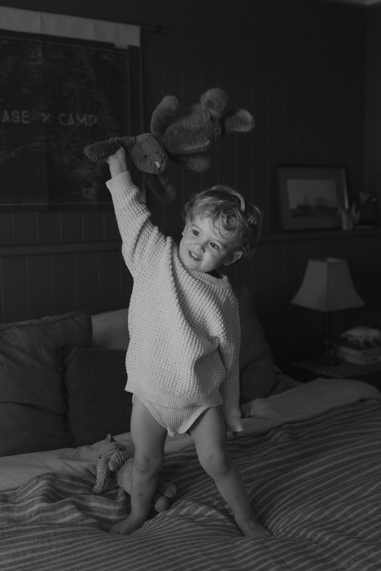 Small child in white sweater playing joyfully with a stuffed animal on a bed in black and white.