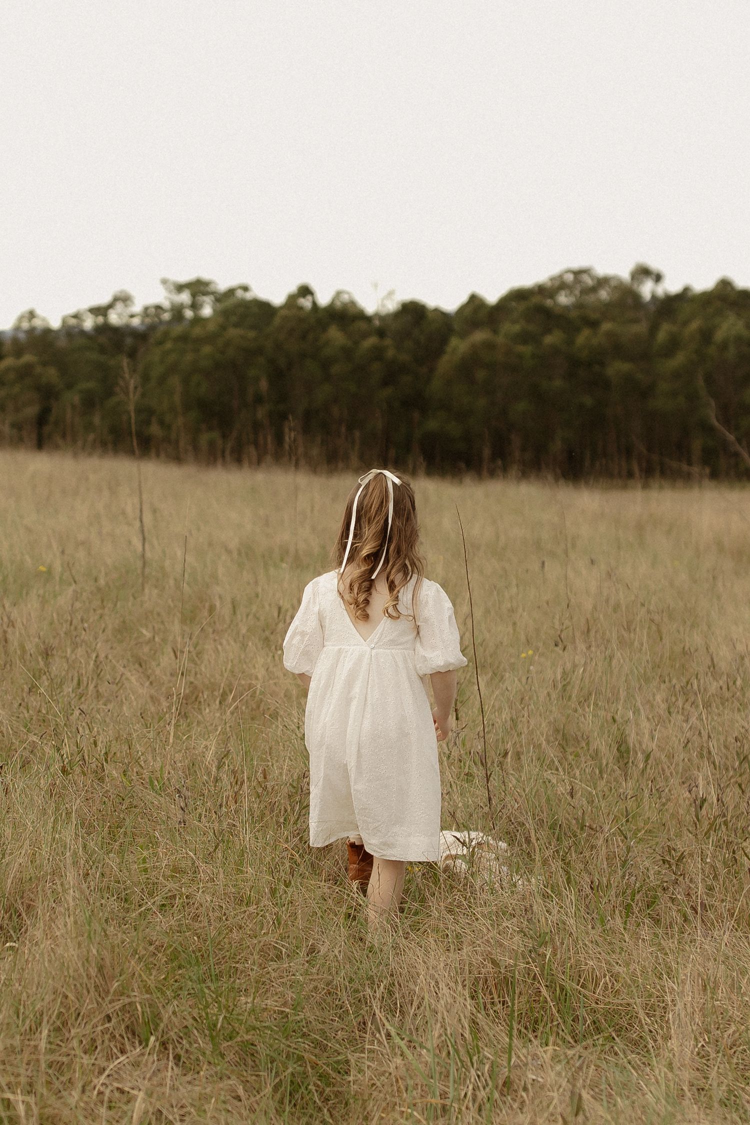 A figure in a flowing white dress walks through a golden field with trees in the distant background.