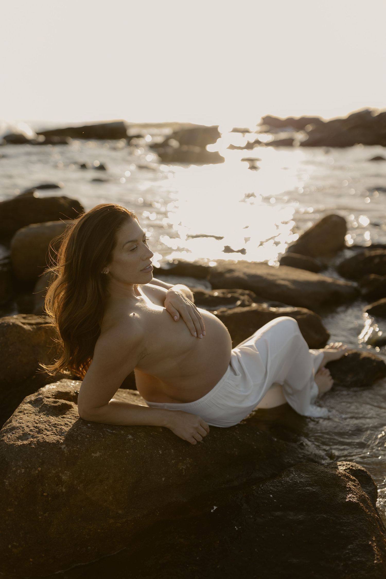 Expectant mother in white dress reclining on coastal rocks at golden hour.
