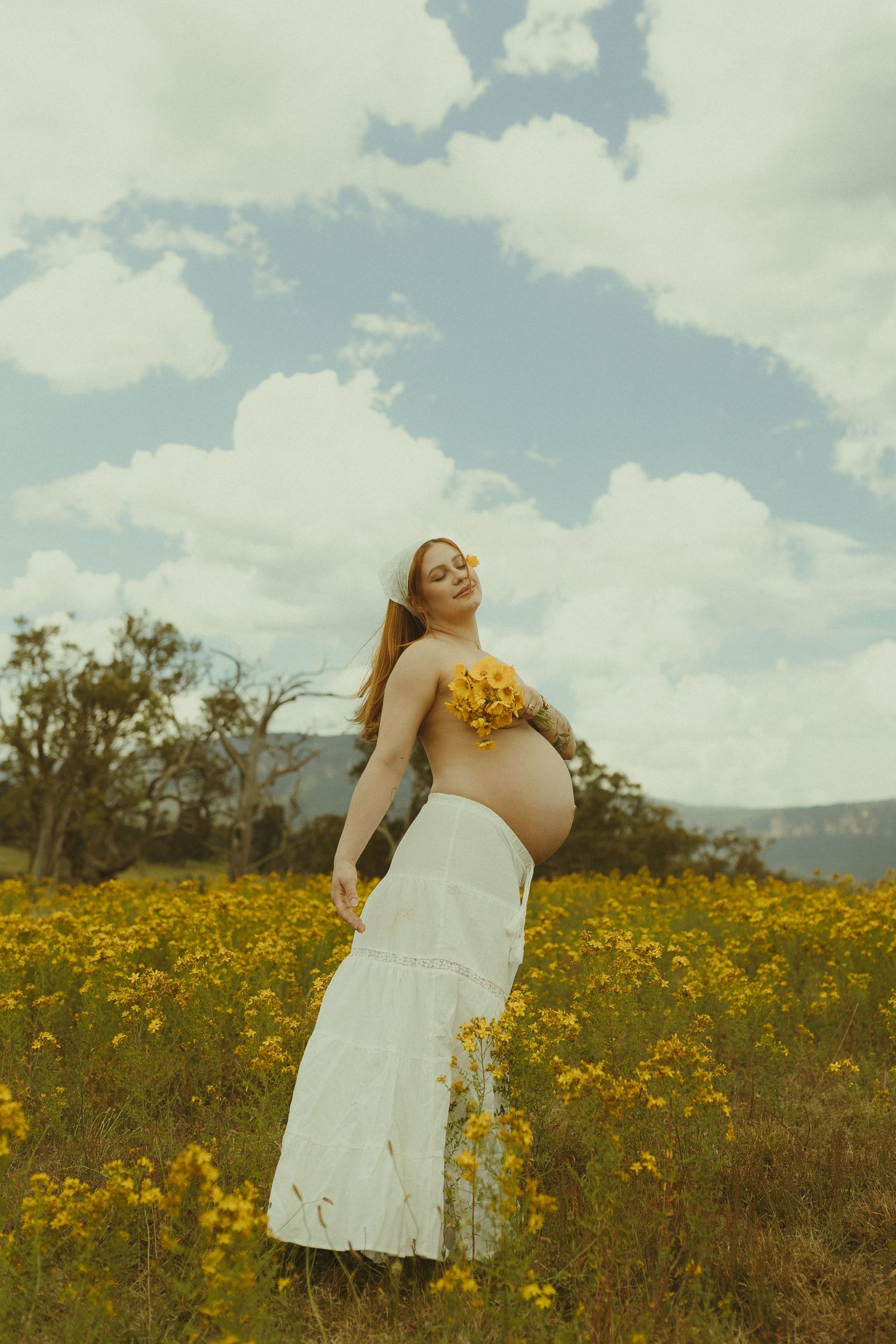 A person in white holds yellow flowers while standing in a sun-drenched meadow under blue skies.