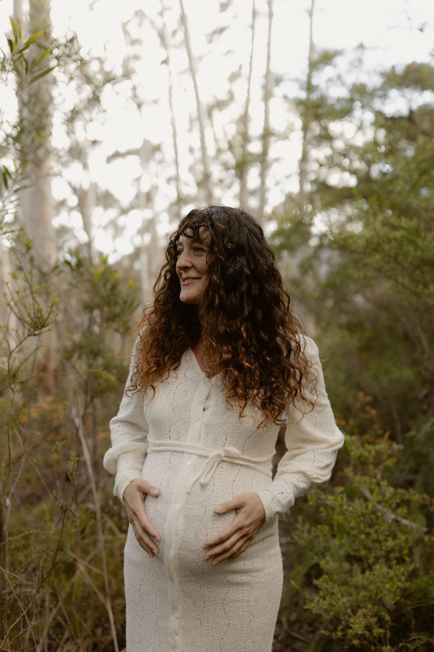 A person in a white sweater dress stands in nature, cradling their pregnant belly while smiling.
