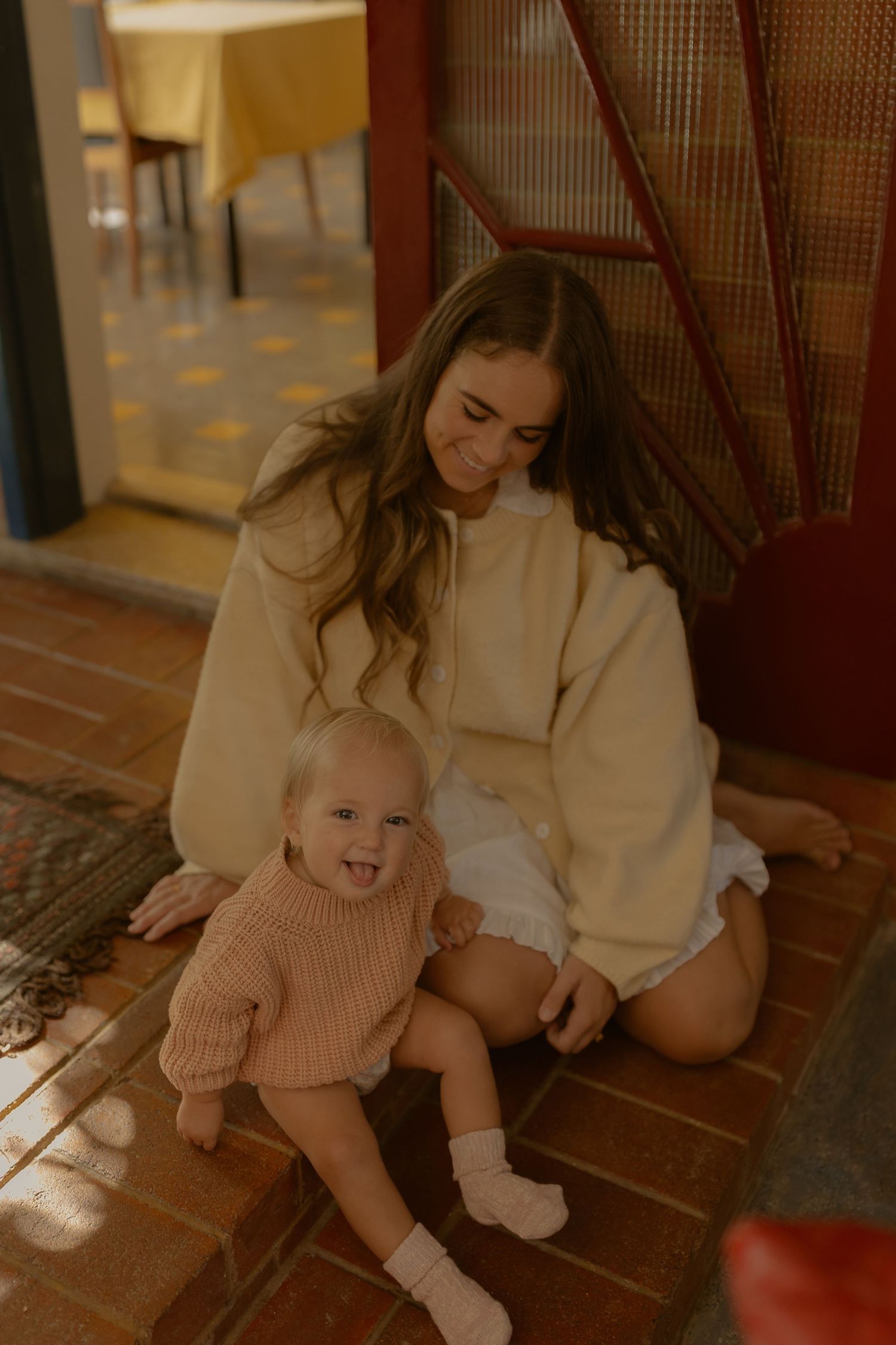 Young woman in cream sweater sits on tiled floor smiling at happy baby in pink knit sweater.