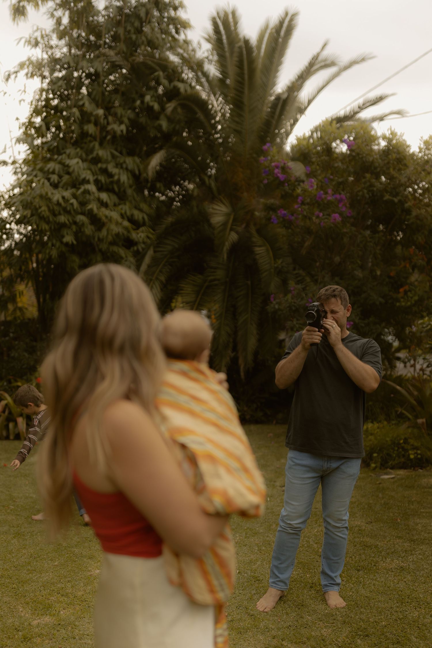 A man photographs a woman holding a baby in a tropical garden setting on an overcast day.
