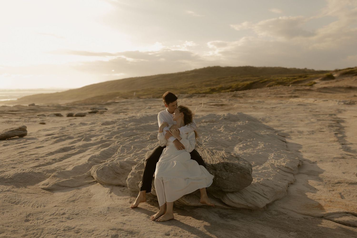 Couple dancing together on rocky beach during golden sunset with dramatic cloudy sky.