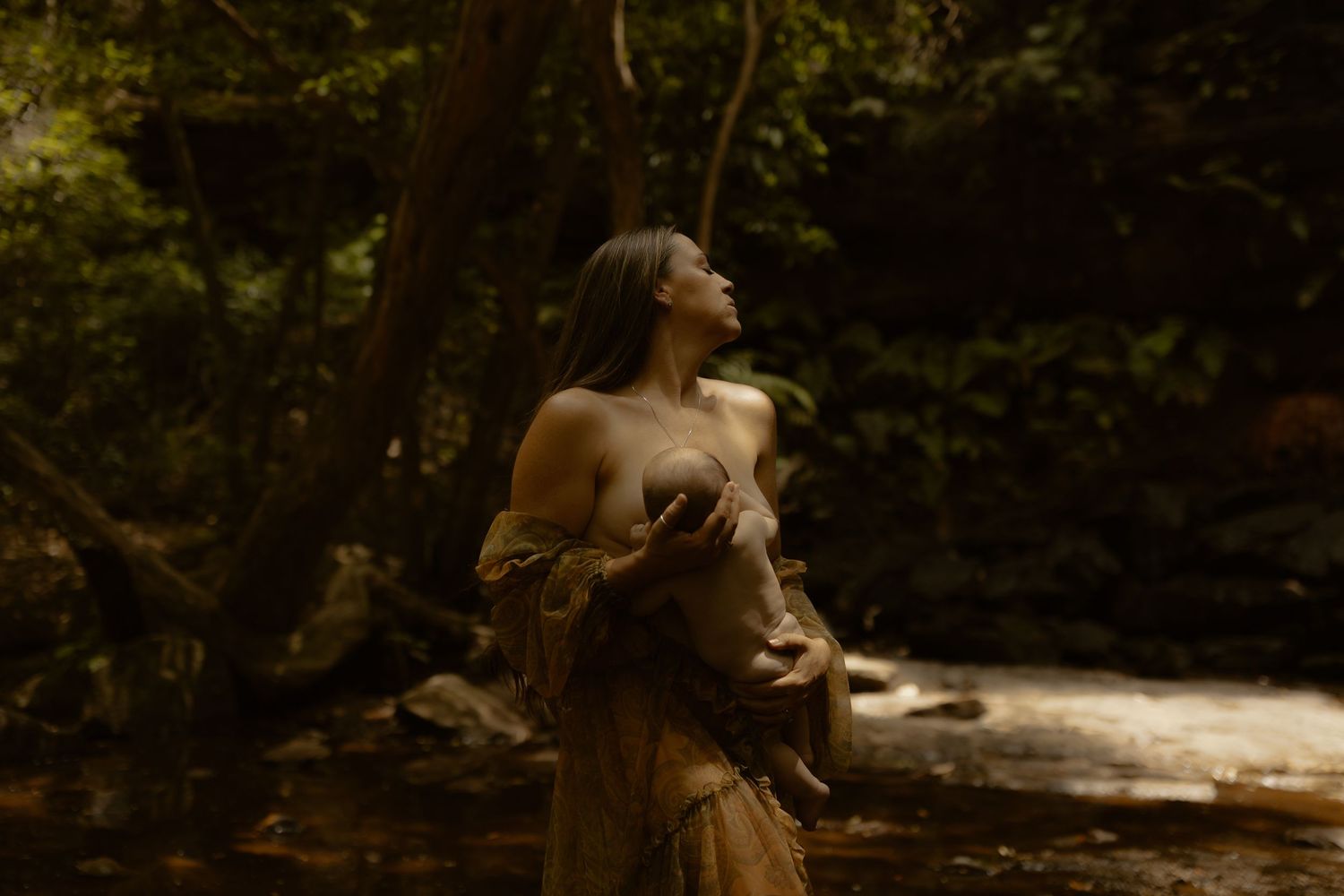 A figure in a flowing dress stands in a dreamy forest setting with sunlight filtering through trees near a stream.