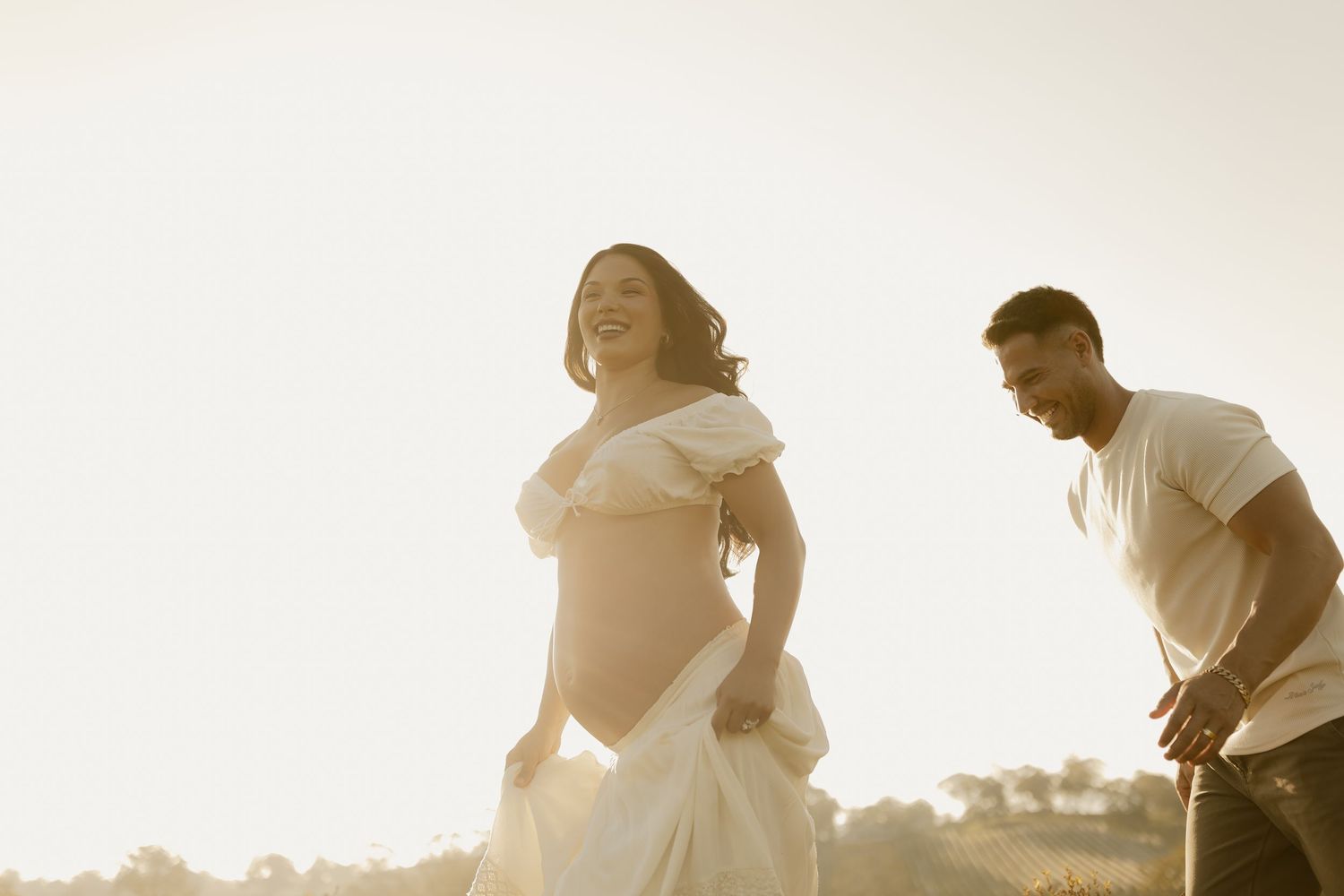 Couple sharing a joyful moment in a sun-drenched field during a maternity photoshoot.