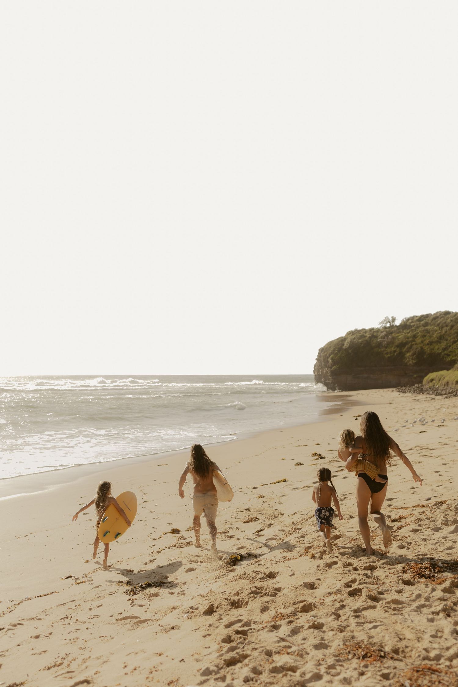 A group walks along a scenic beach carrying surfboards during a hazy afternoon.
