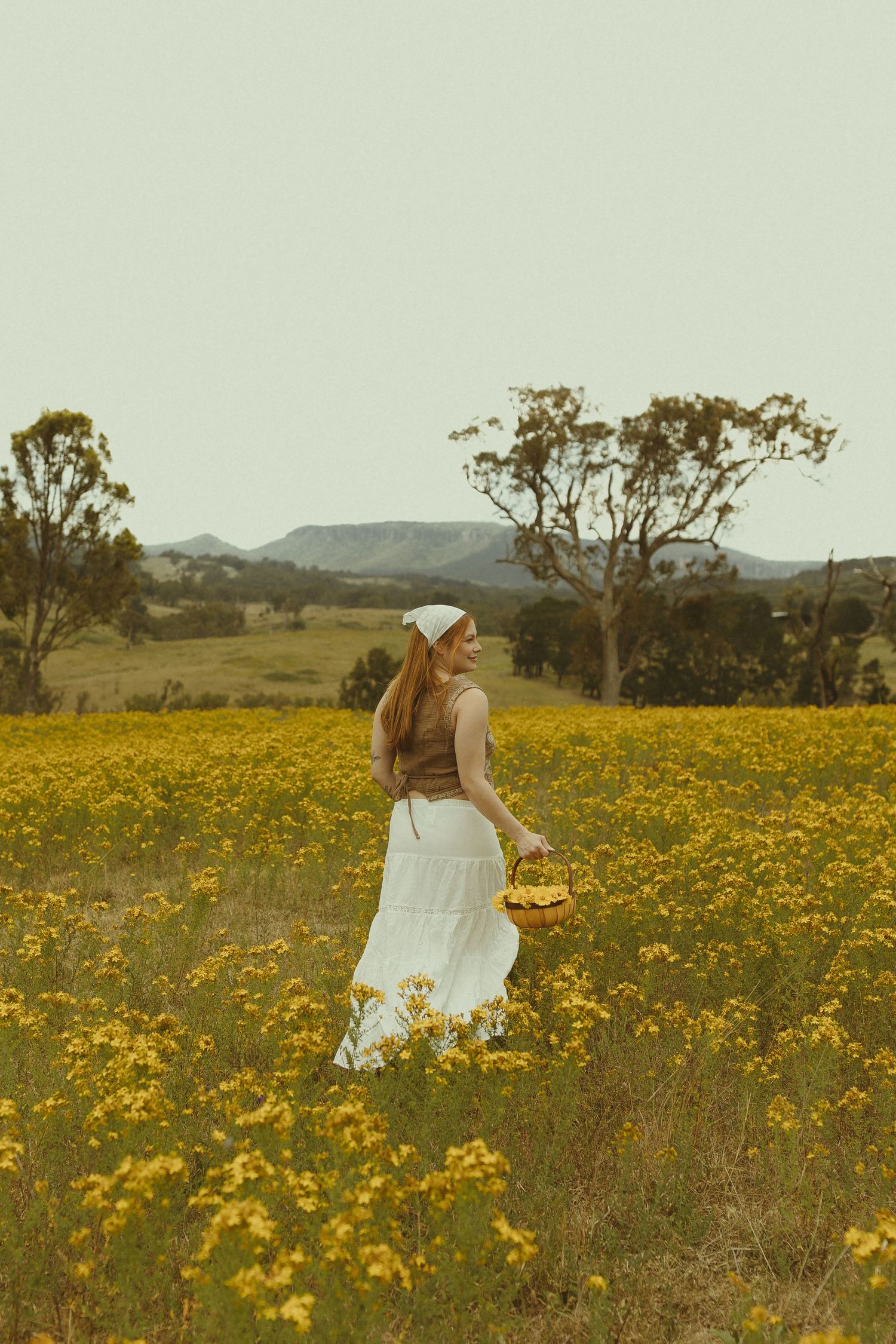 A dreamy scene of someone in a white dress walking through a vibrant field of yellow wildflowers with mountains in the distance.