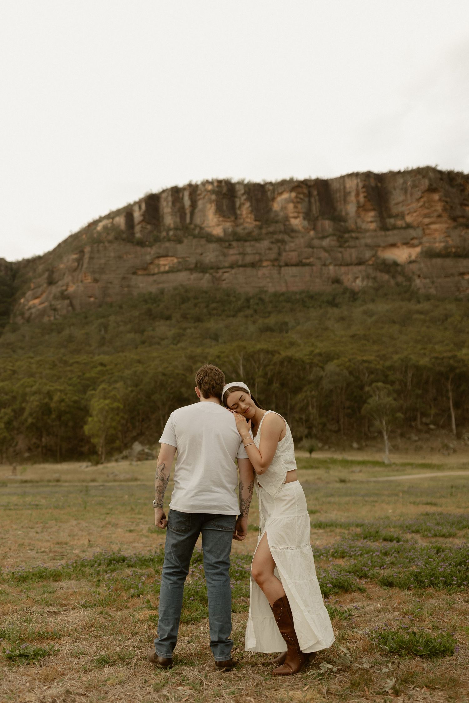 A couple in casual attire stands together in a field with dramatic cliffs and rock formations in the background.