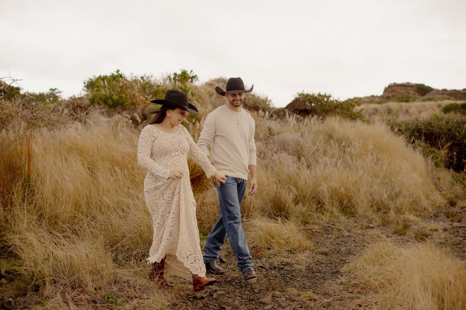 A couple walks together through tall golden grass in a scenic desert landscape at sunset.