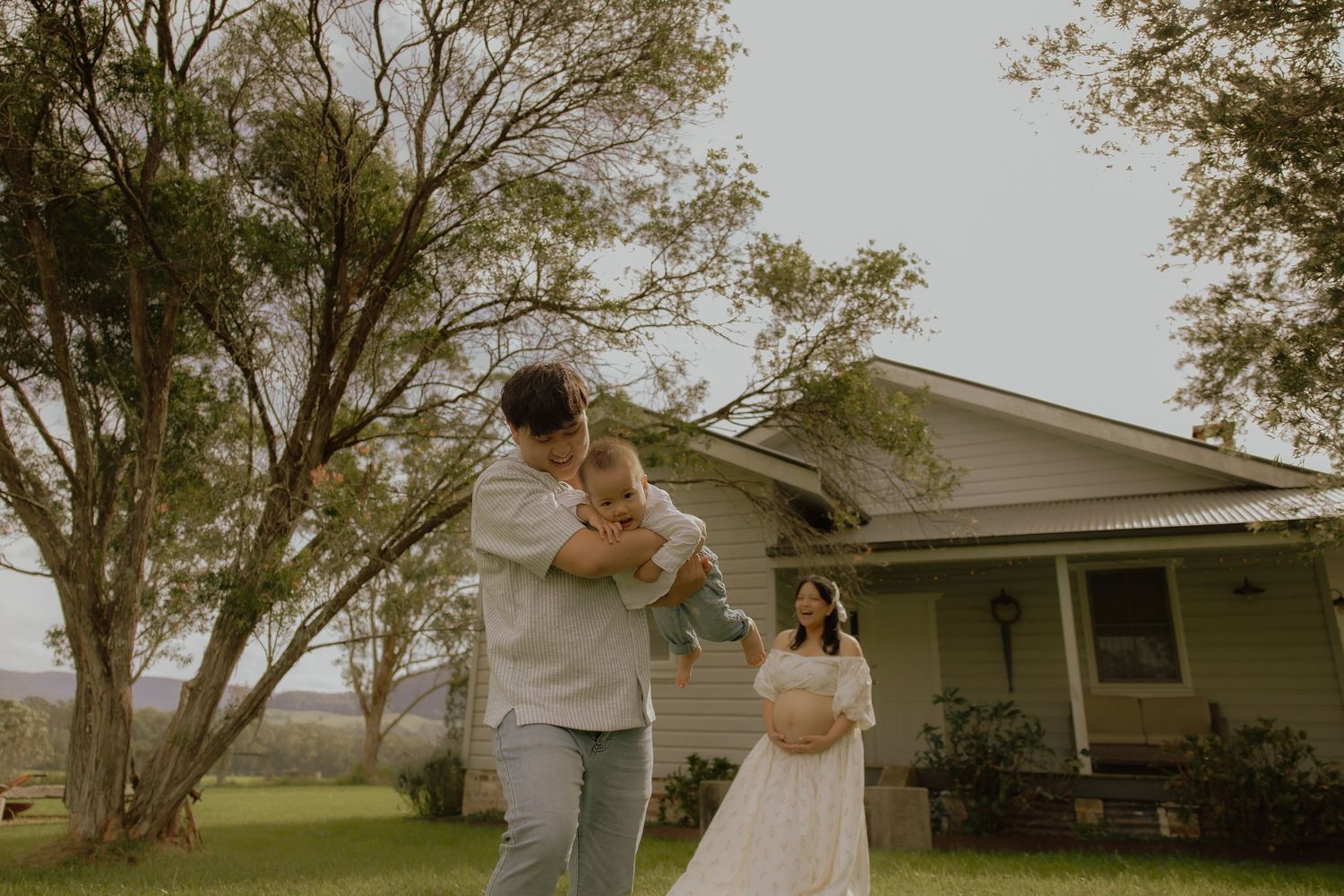 Family of three with baby outside a charming country home surrounded by lush green trees.