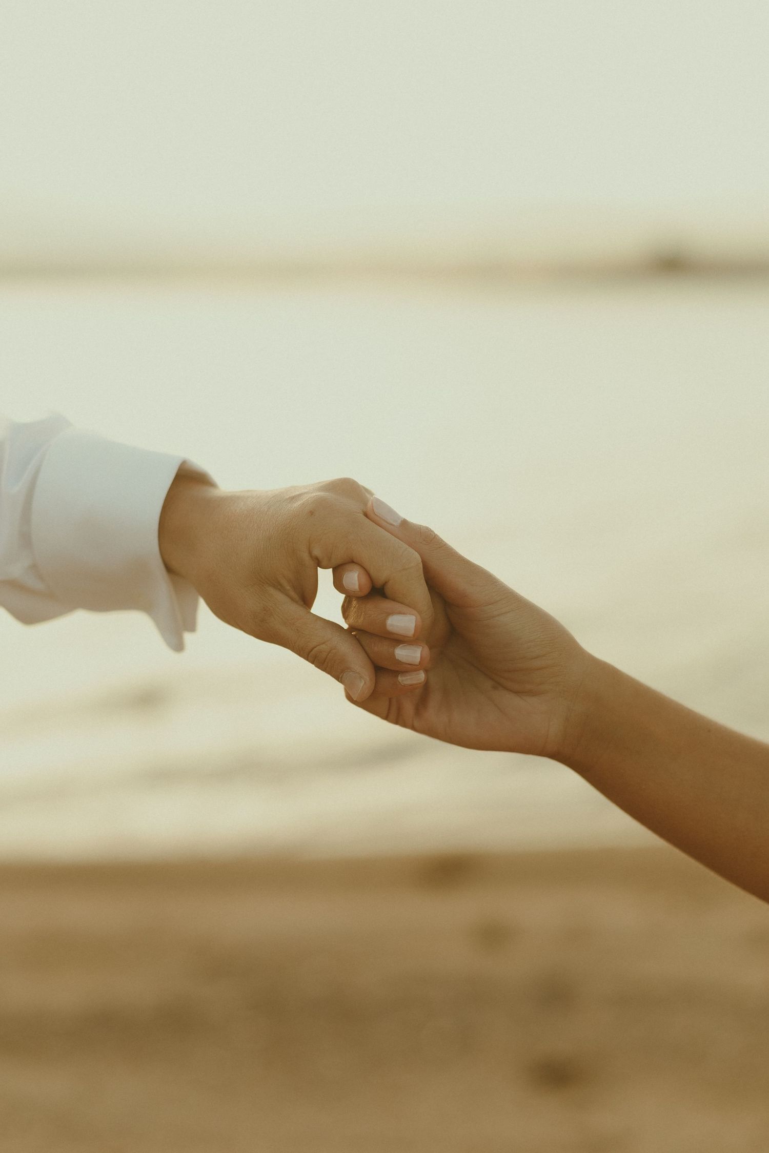 Two hands tenderly reaching for each other against a soft, blurred beach background in warm light.