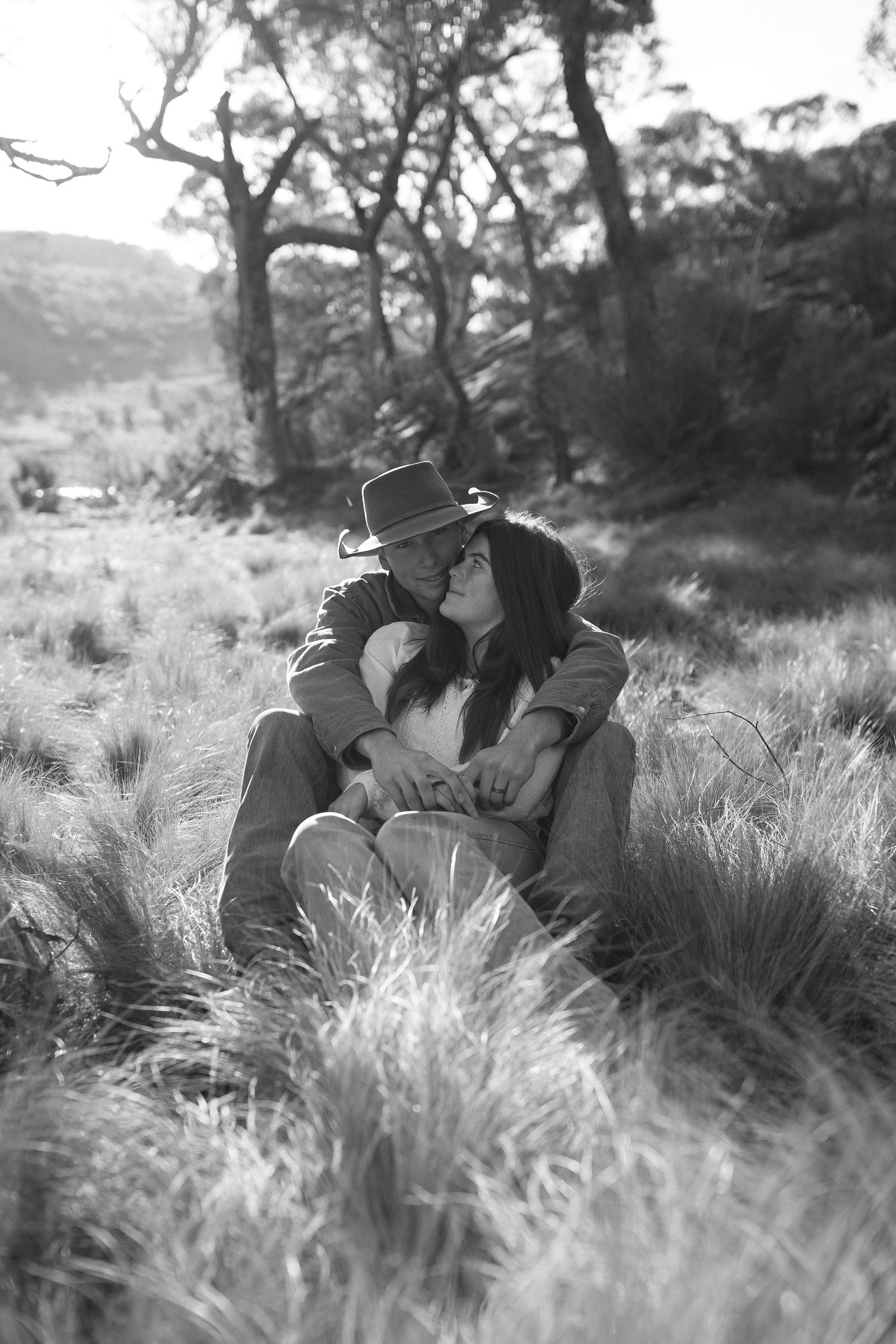 A romantic black and white photograph of a couple embracing in tall grass with trees in the background.