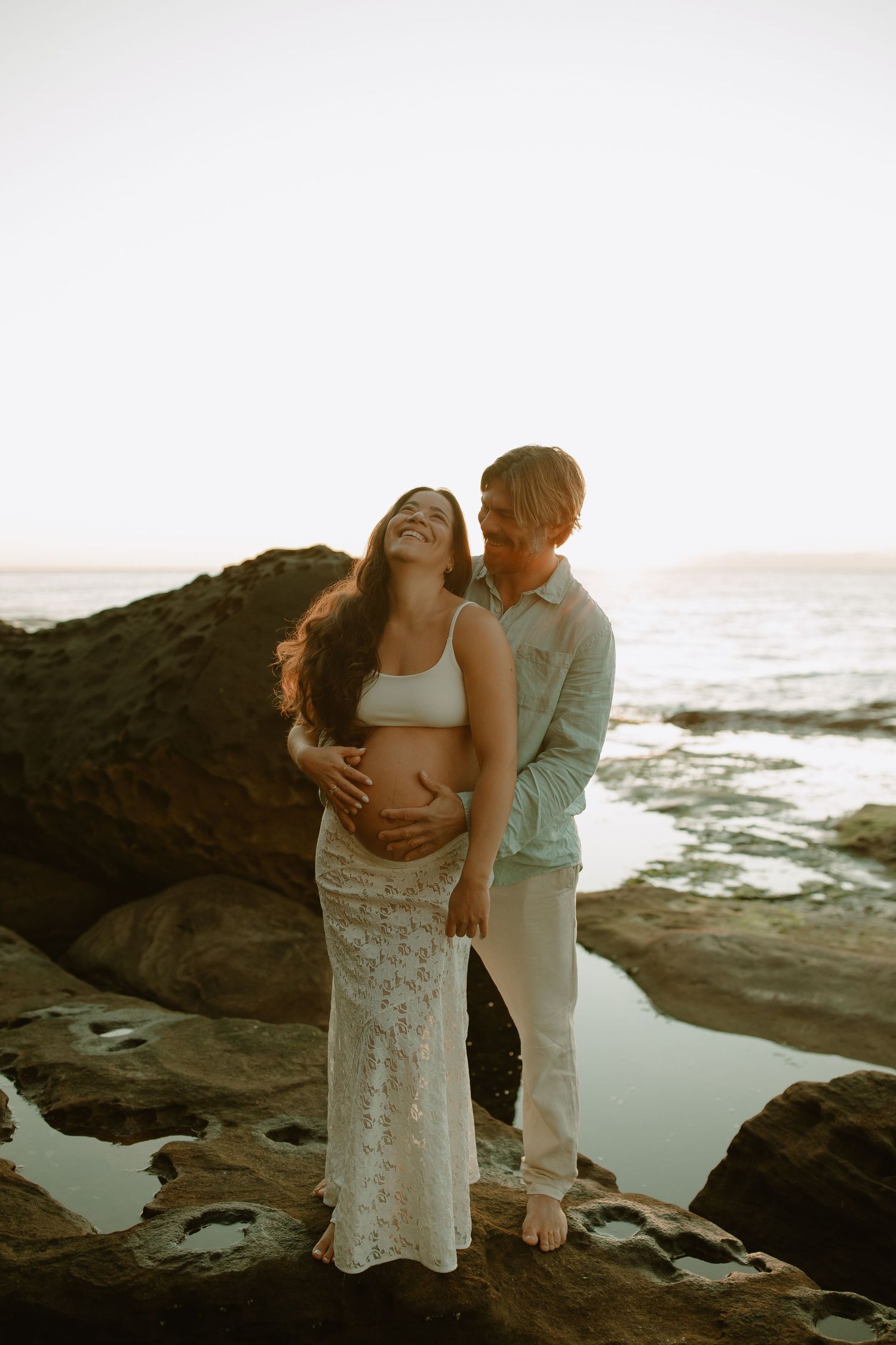 Couple embraces intimately on rocky coastal shore during sunset maternity photoshoot.