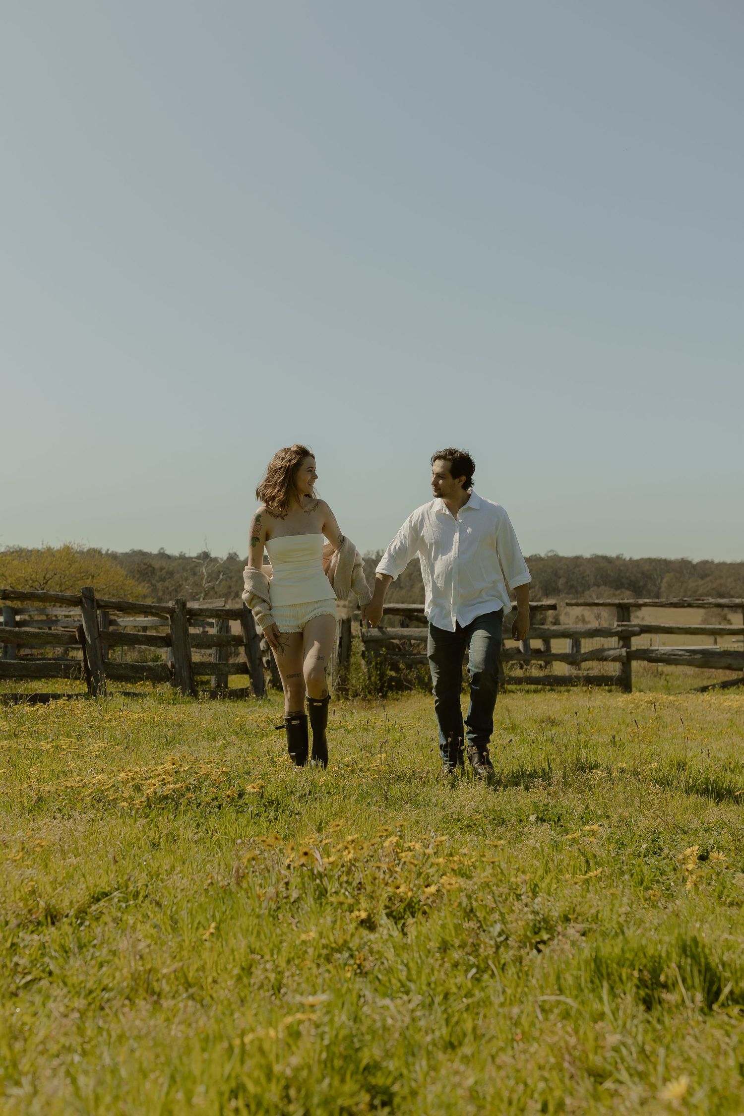 Two people walk hand in hand through a grassy meadow with wildflowers and a rustic fence in the background.