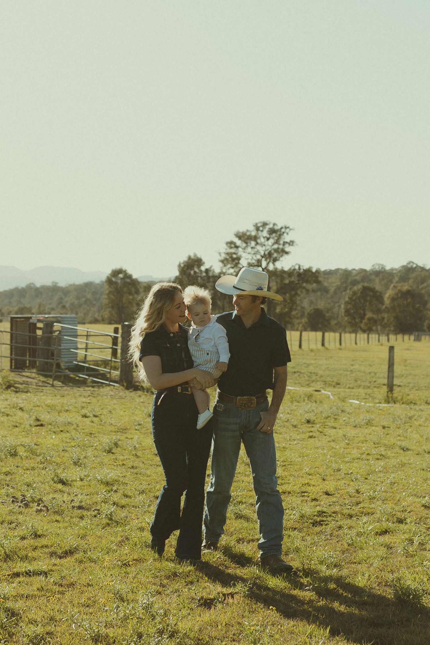 A family stands together in a rustic farm field with fencing and trees in the background.