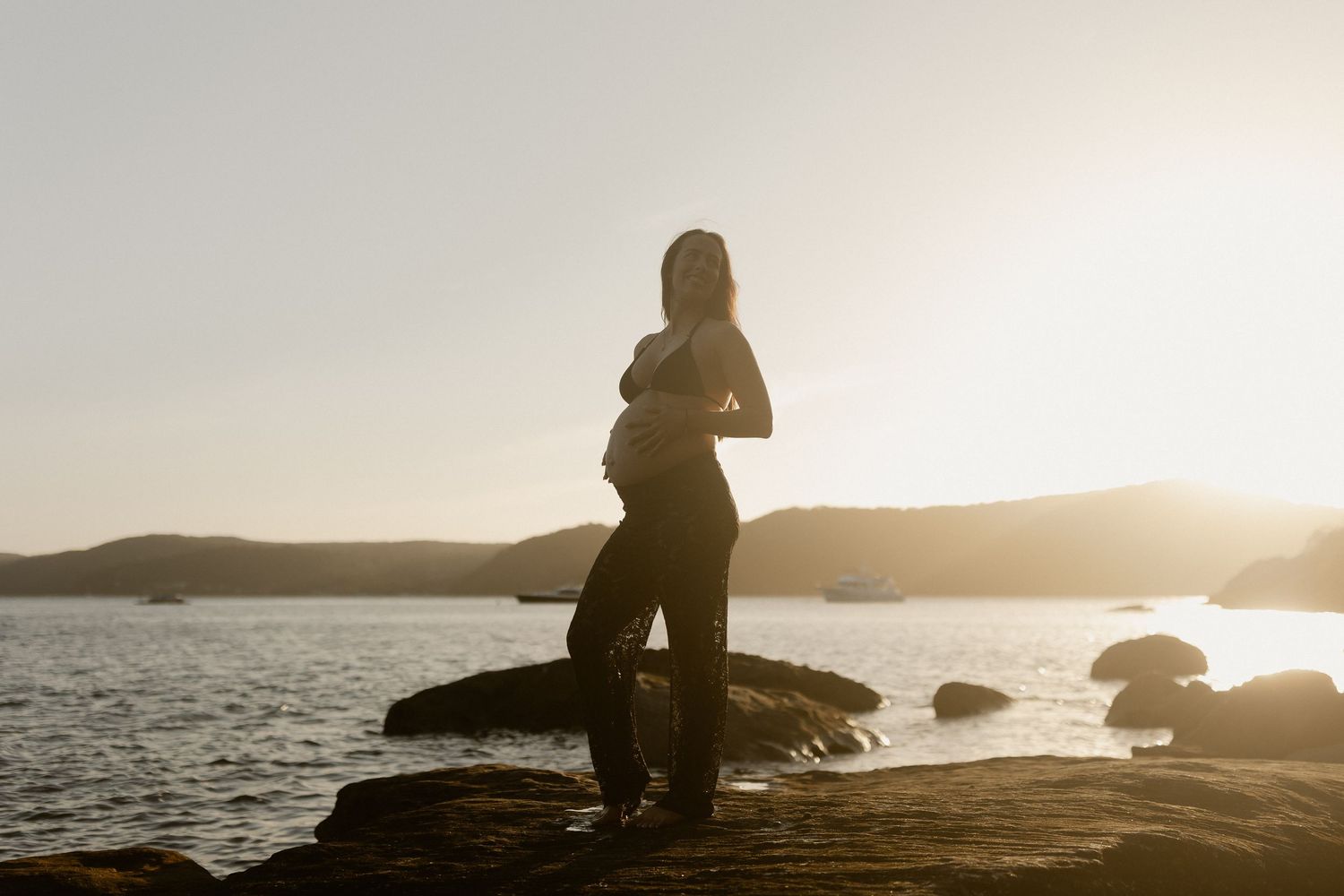 Silhouetted figure sitting by calm ocean at sunset with warm golden light reflecting on water.