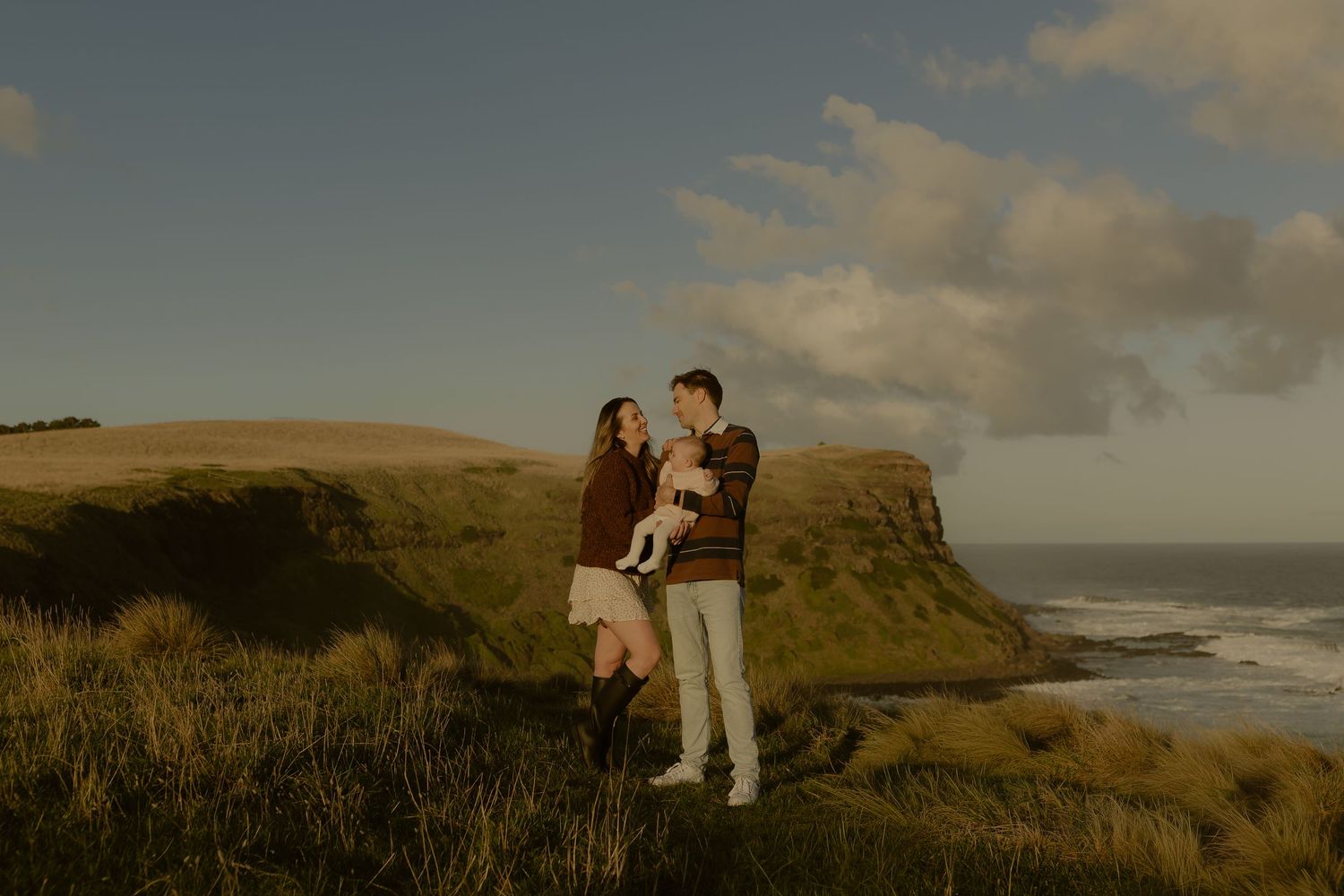 A young family with a baby stands on a coastal cliff surrounded by green hills and ocean scenery.