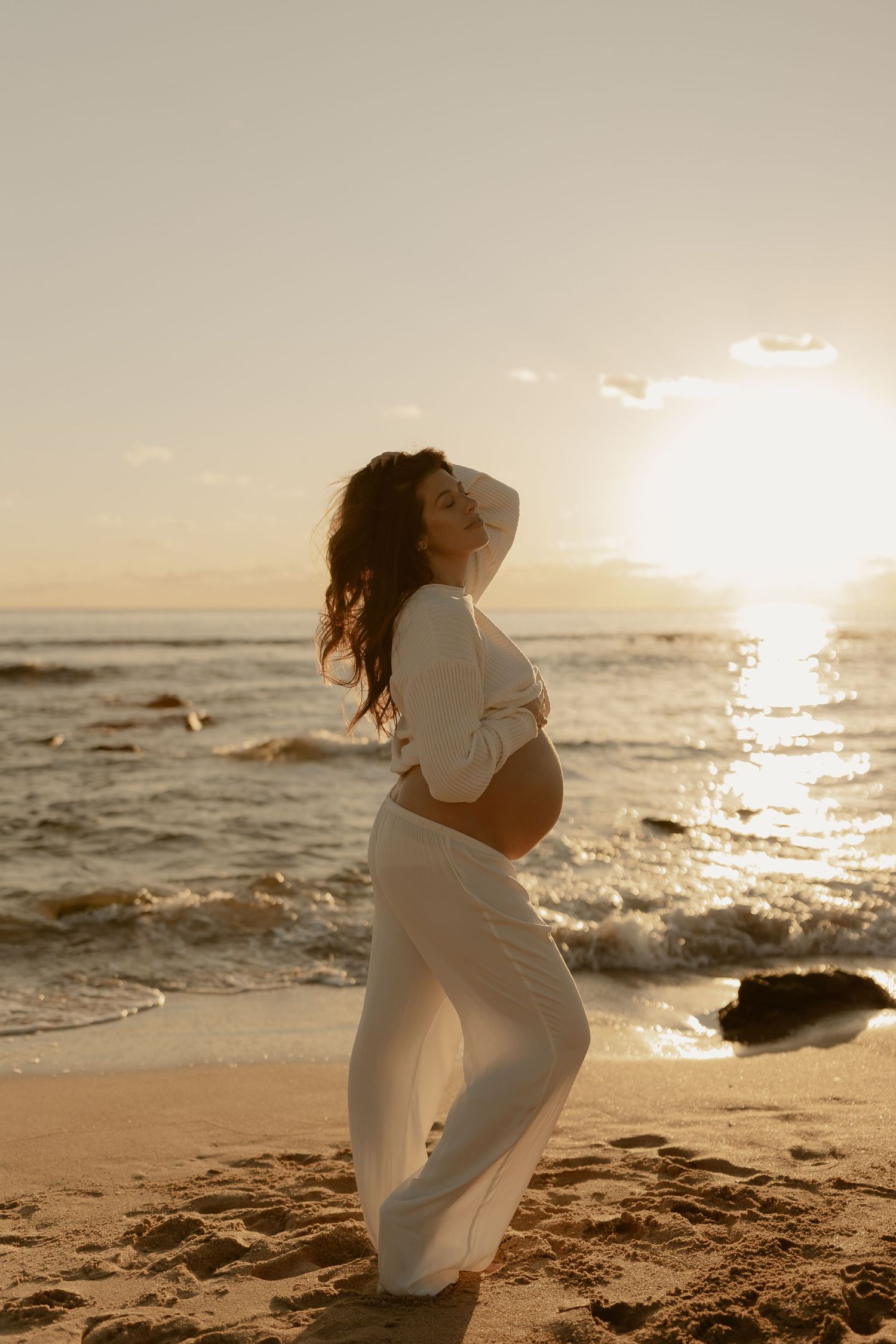 Pregnant woman in white outfit posing on beach at sunset, holding belly with ocean waves behind.