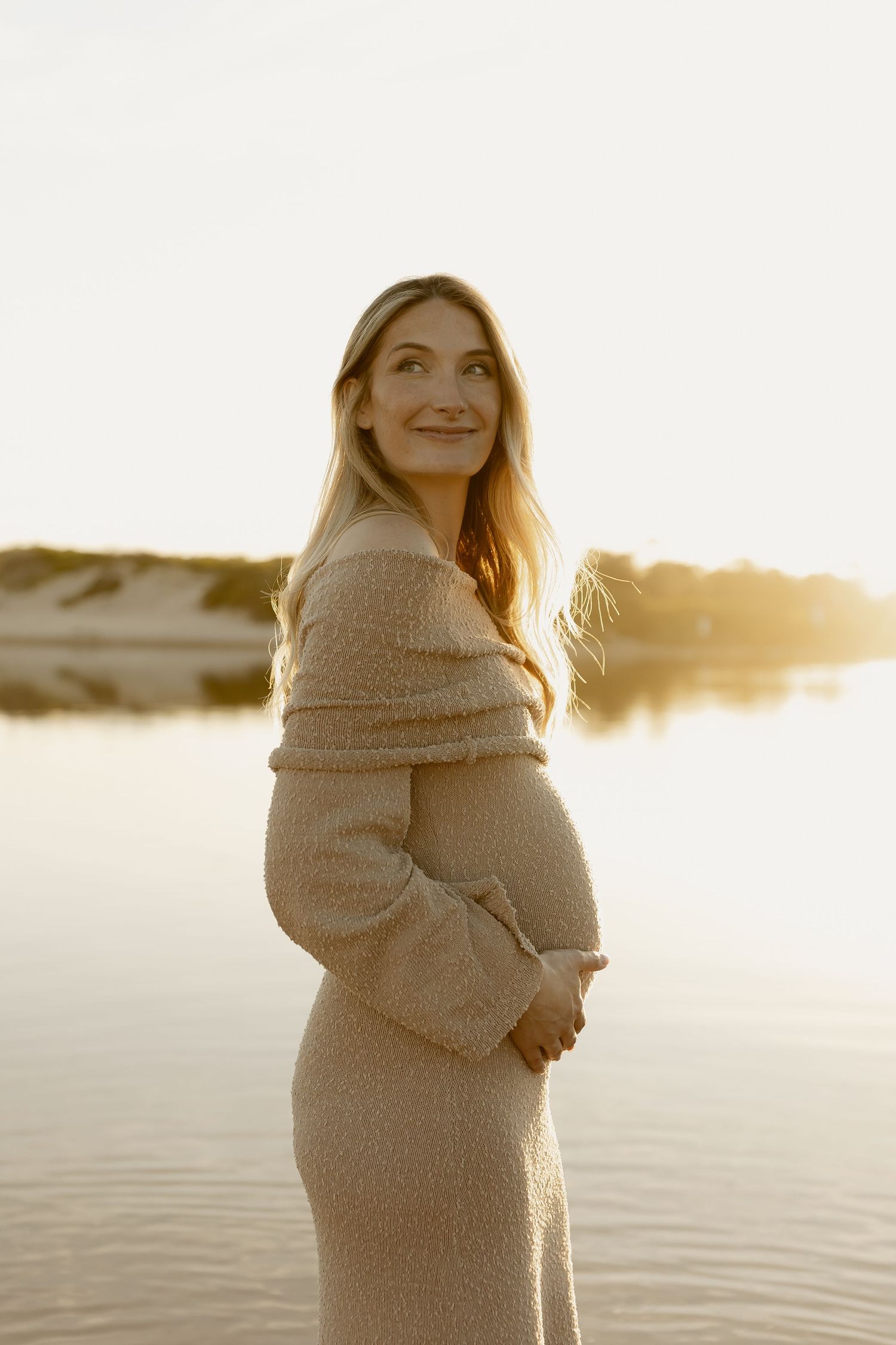 A pregnant woman in a beige knit off-shoulder dress cradles her bump at golden hour by a lake.