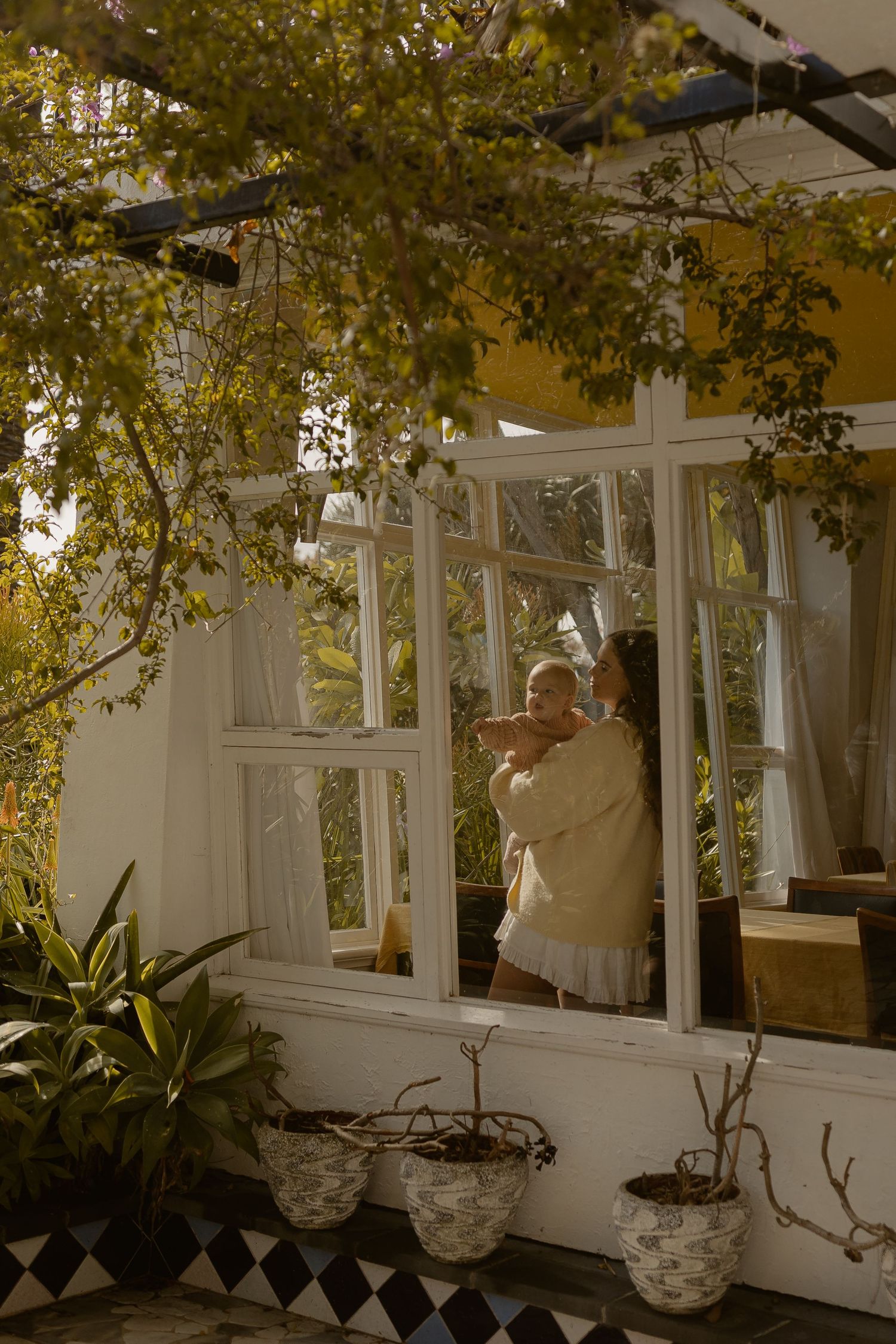 A mother holds her baby near sunlit white-framed windows surrounded by lush green plants and hanging vines.