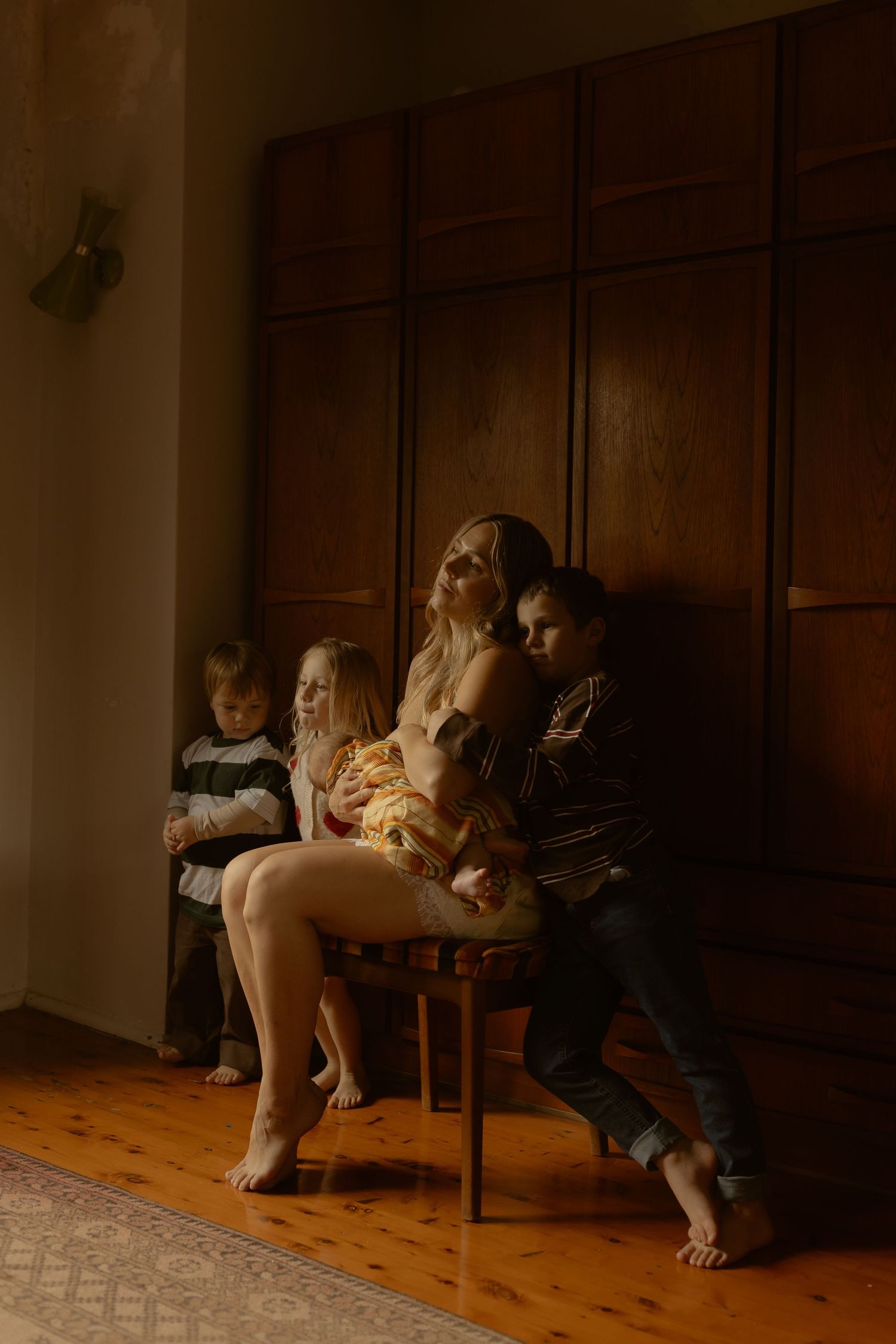 A woman and man sit closely together with three children in a dimly lit room with dark wooden paneling.