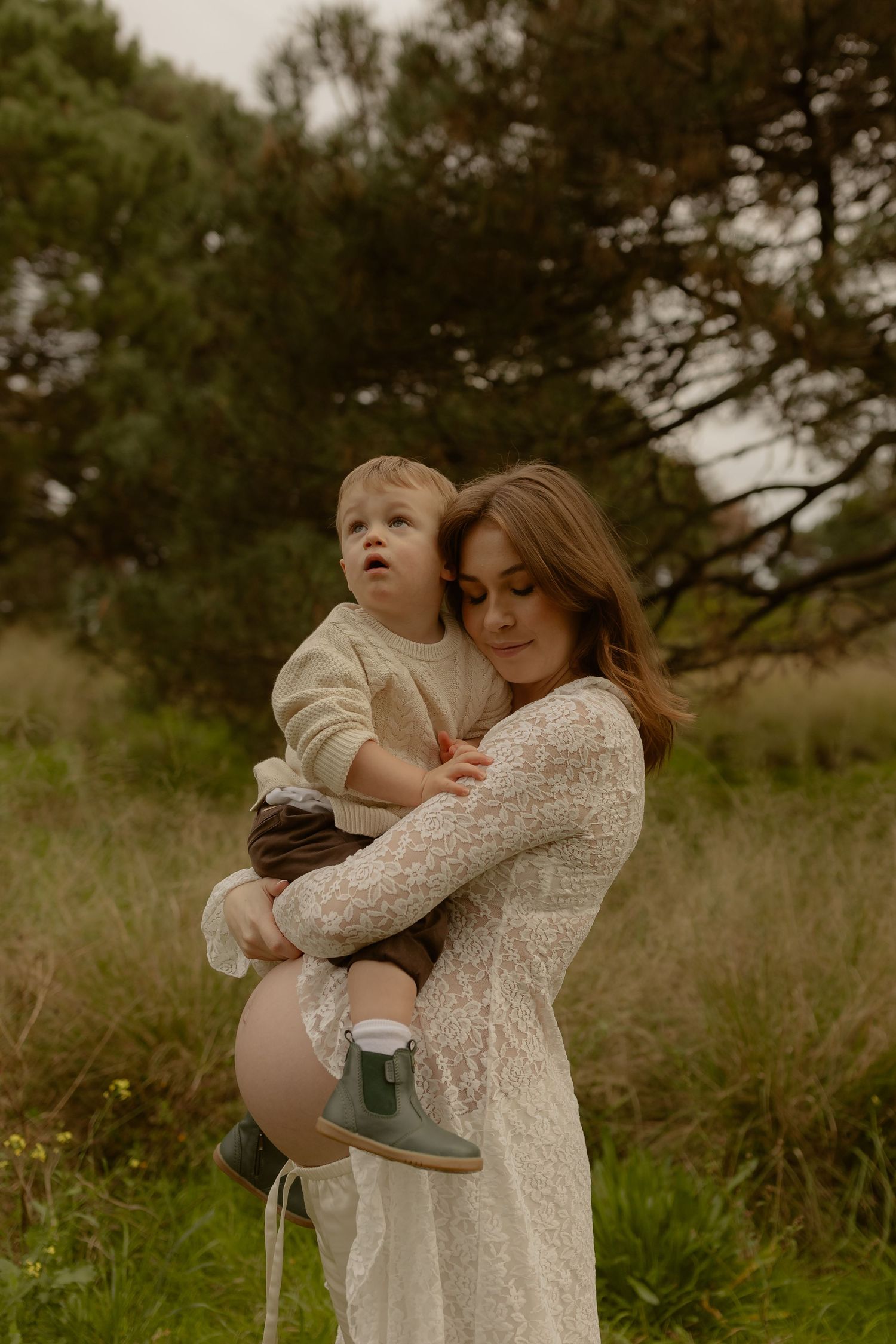 Pregnant woman in white lace dress holding toddler boy in outdoor nature setting.