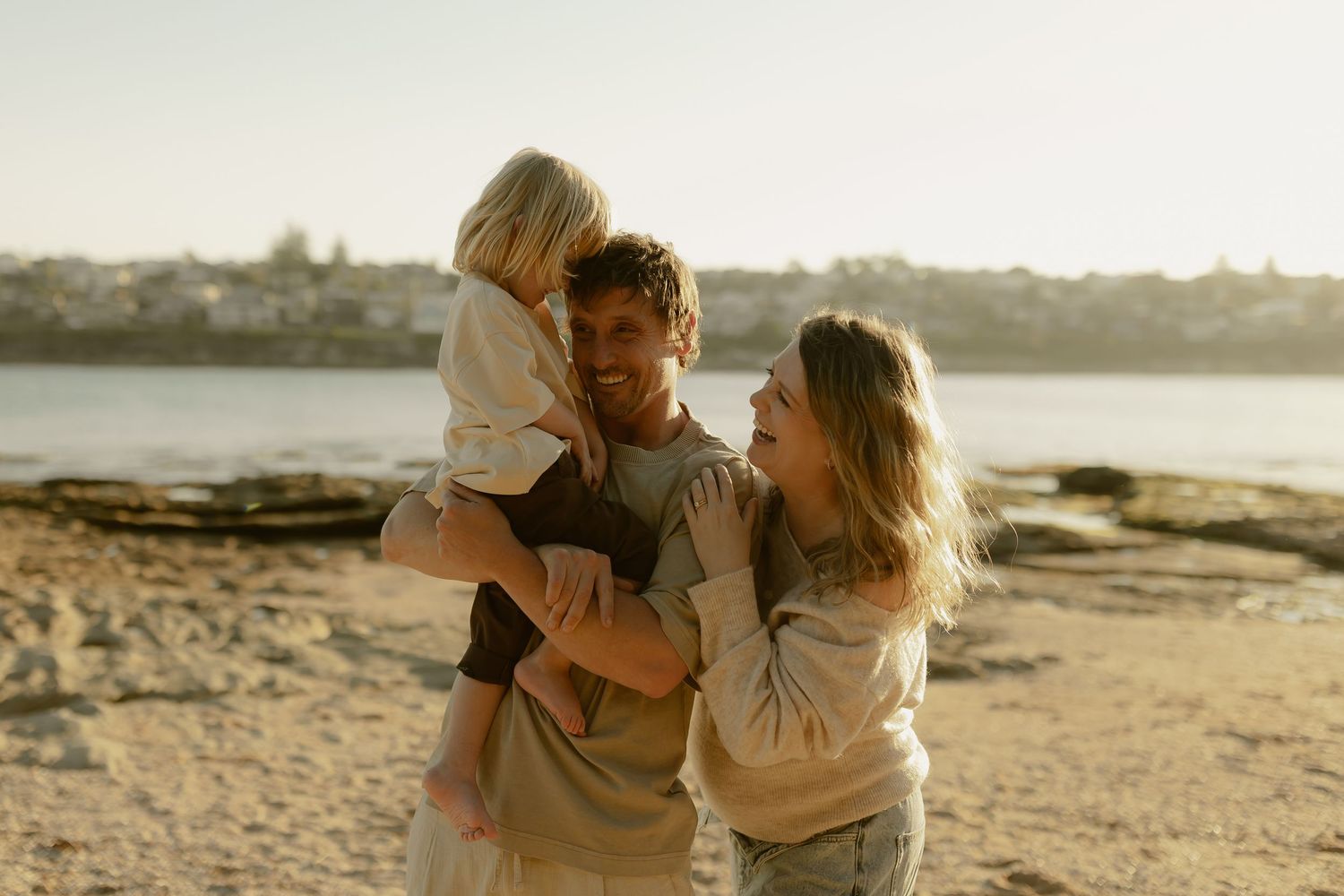 A happy family of three enjoys a sunny beach day, with a man holding a young child while a woman smiles beside them.