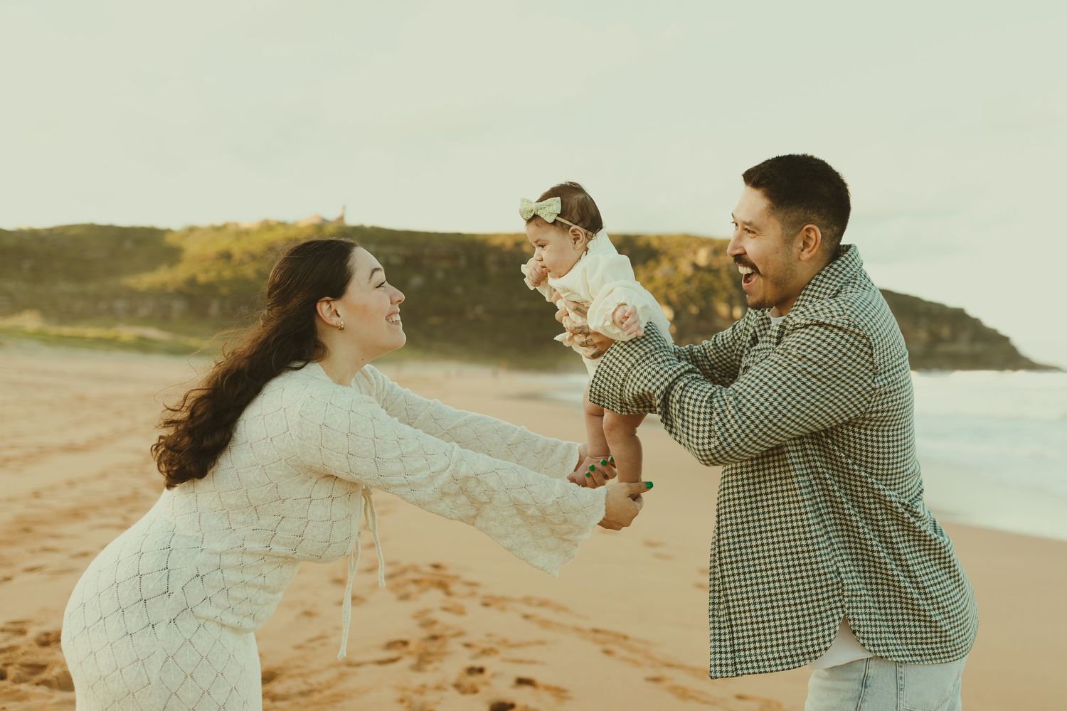 A family enjoys a tender moment together on a sandy beach during golden hour with coastal cliffs in the background.