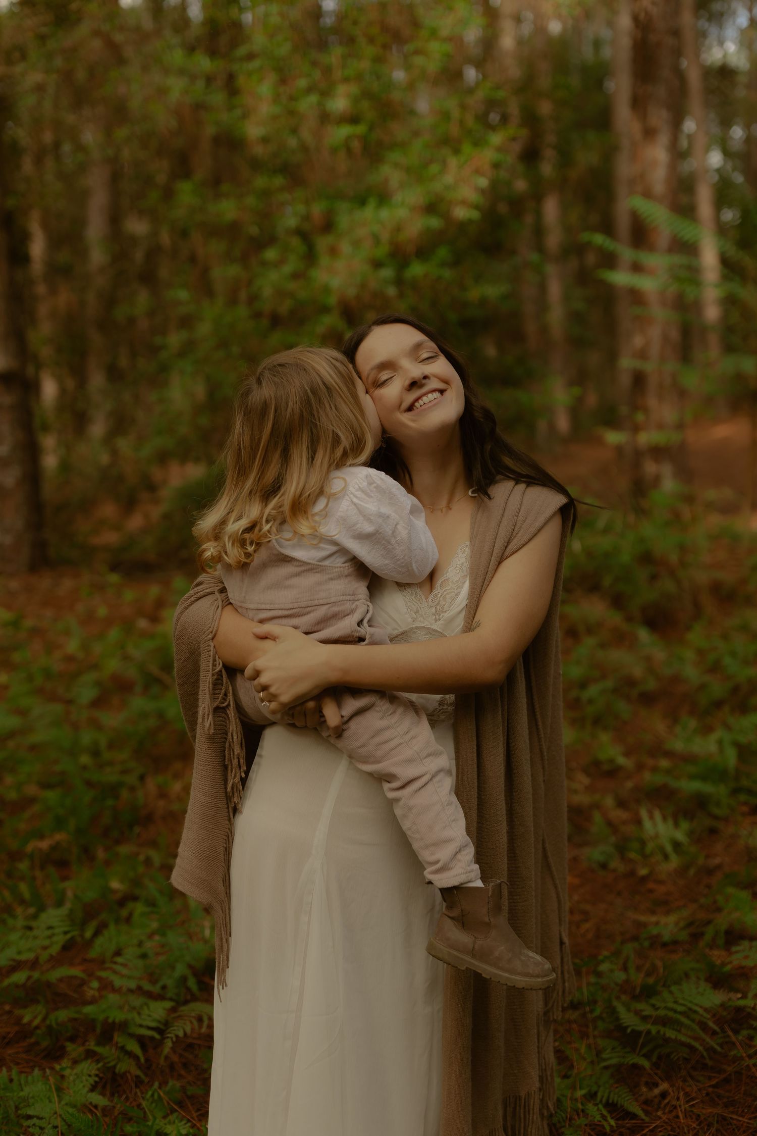Smiling mother in white dress holding young child who kisses her cheek in forest.
