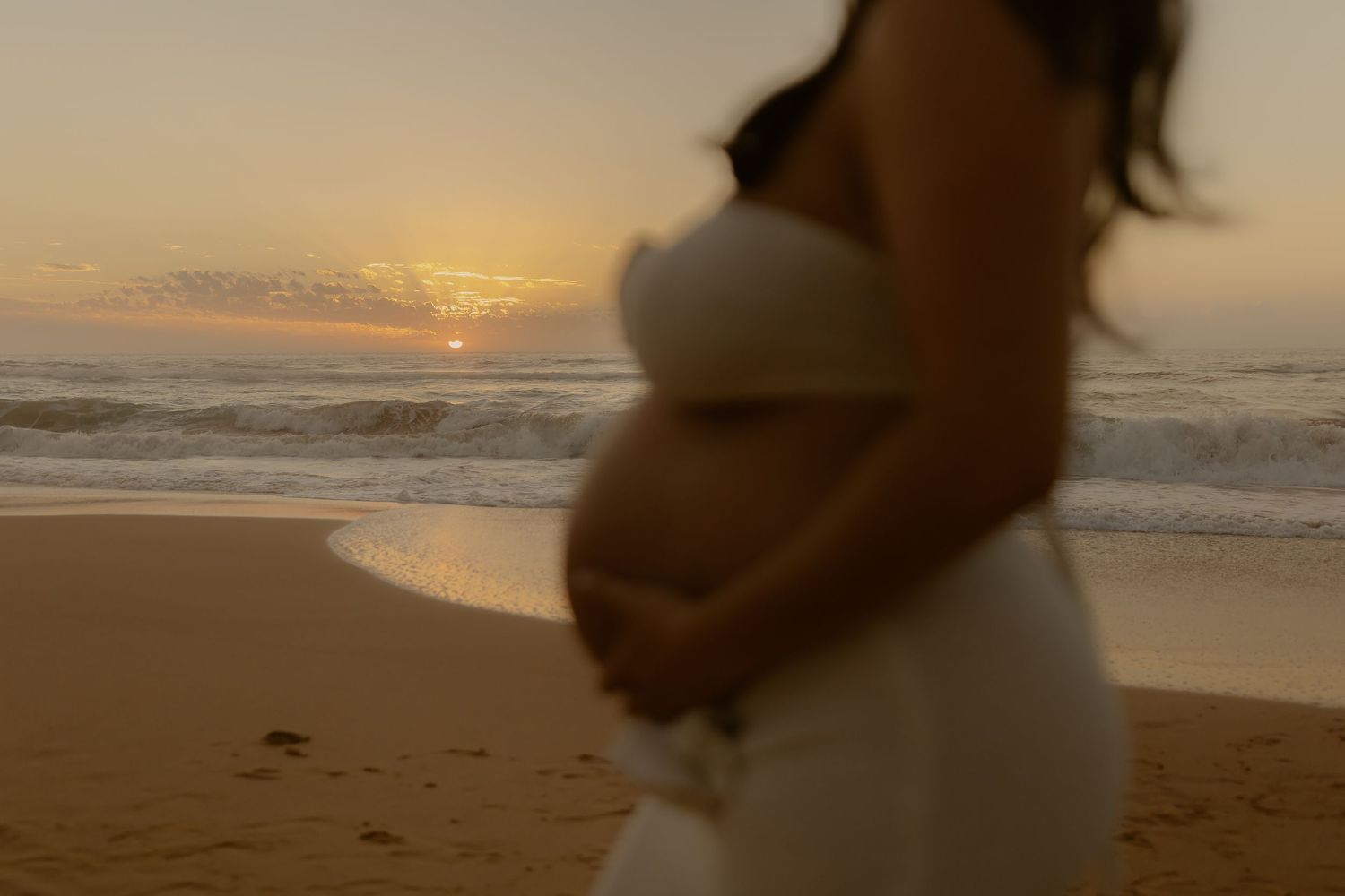 A pregnant silhouette in a white dress stands on a beach at sunset while holding her belly.