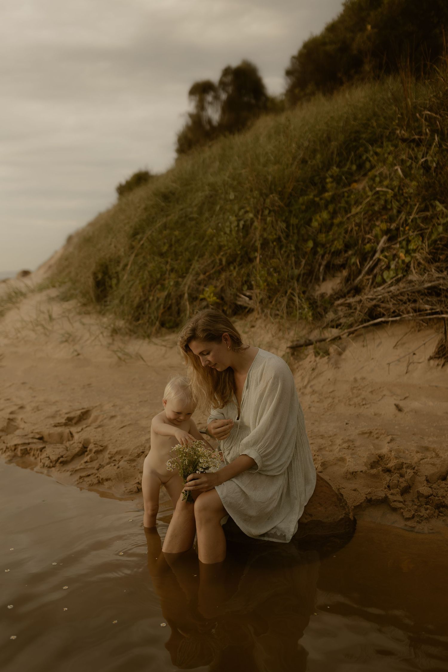 Person sits at the edge of a sandy beach near water during golden hour in a moody atmospheric setting.