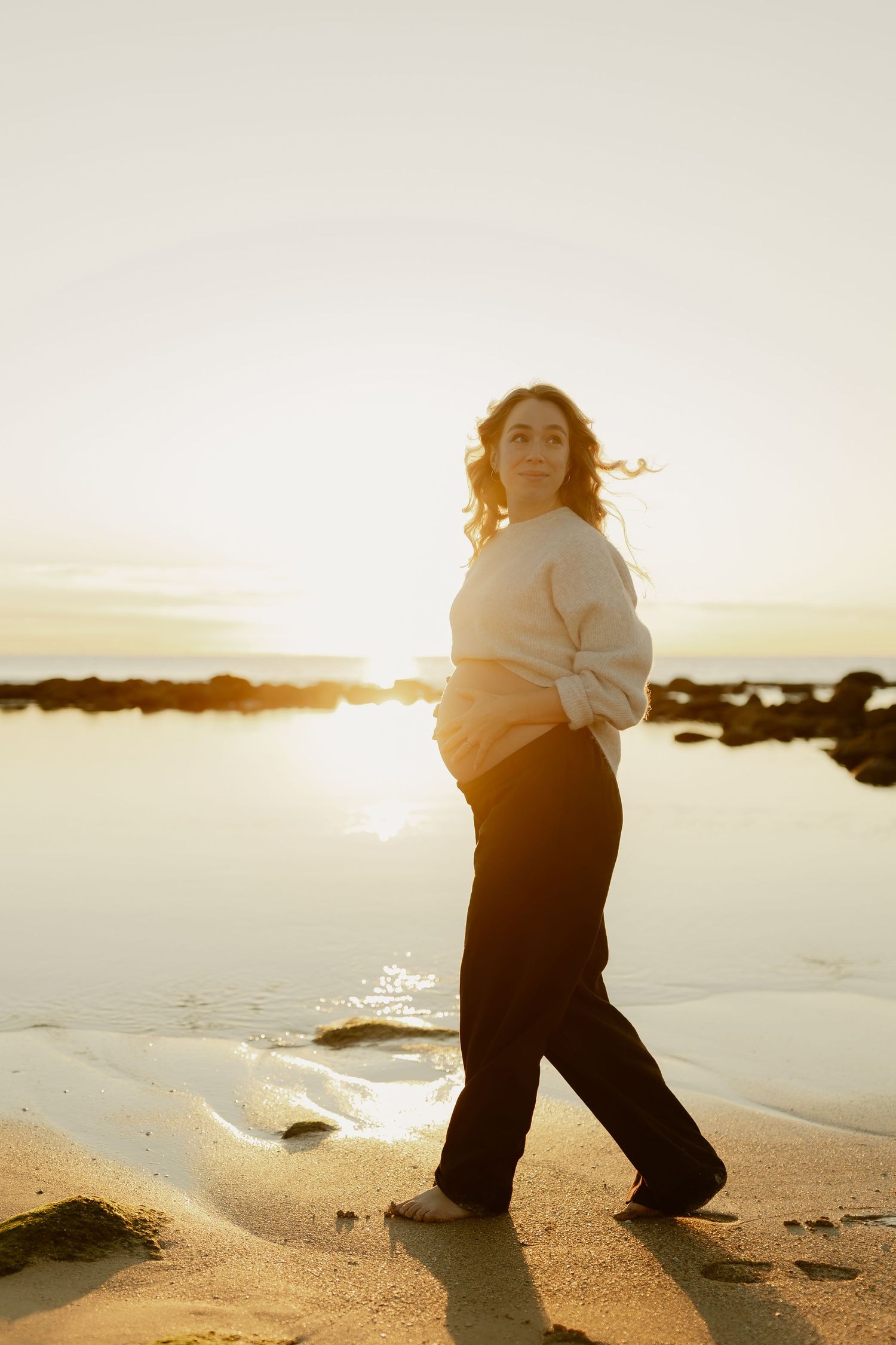 Pregnant woman in cream sweater walks along beach at sunset, cradling baby bump in golden light.