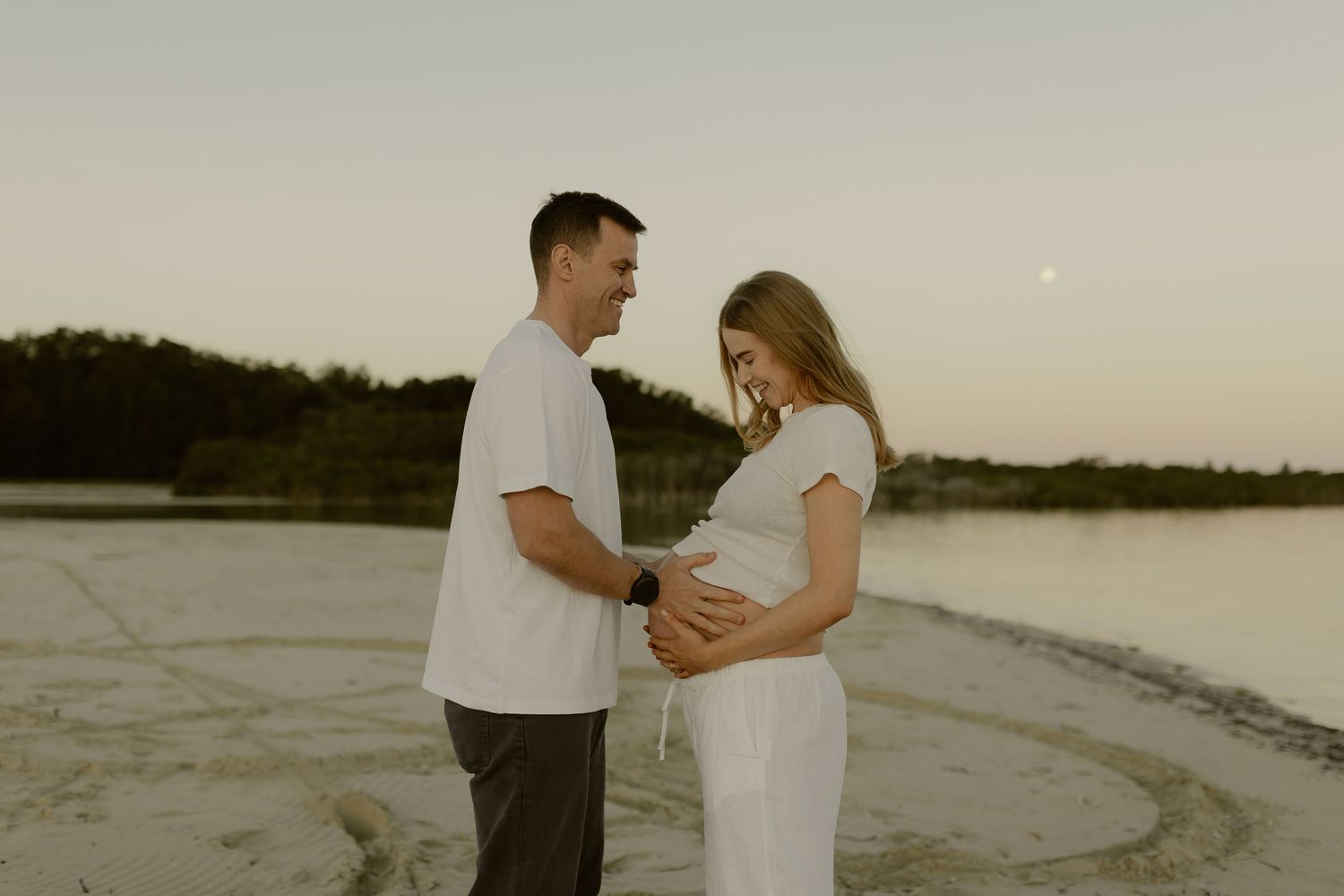 Couple sharing tender moment on beach at dusk, woman in white dress showing baby bump.