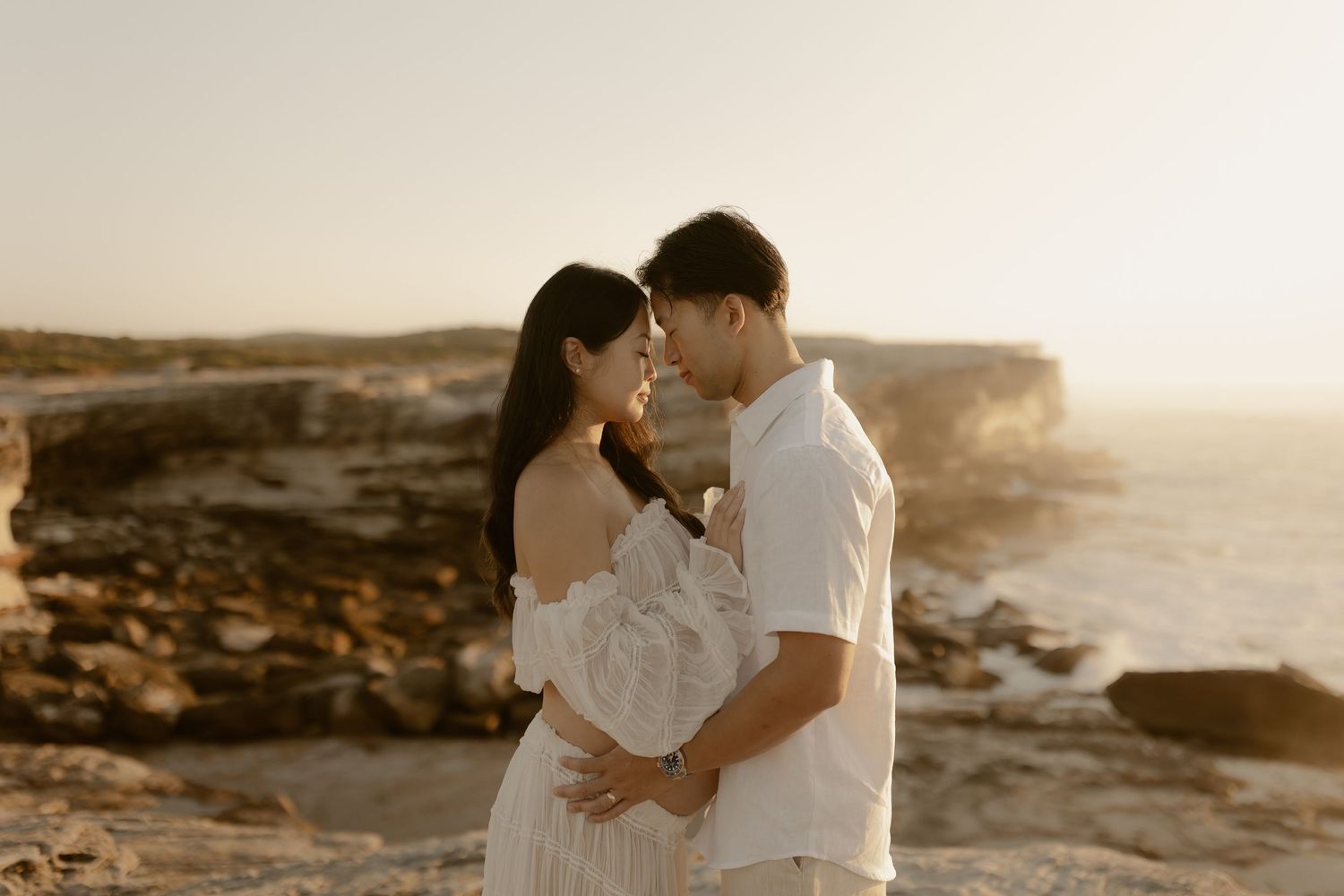 Romantic couple embraces during golden hour sunset at a scenic coastal cliff overlook.