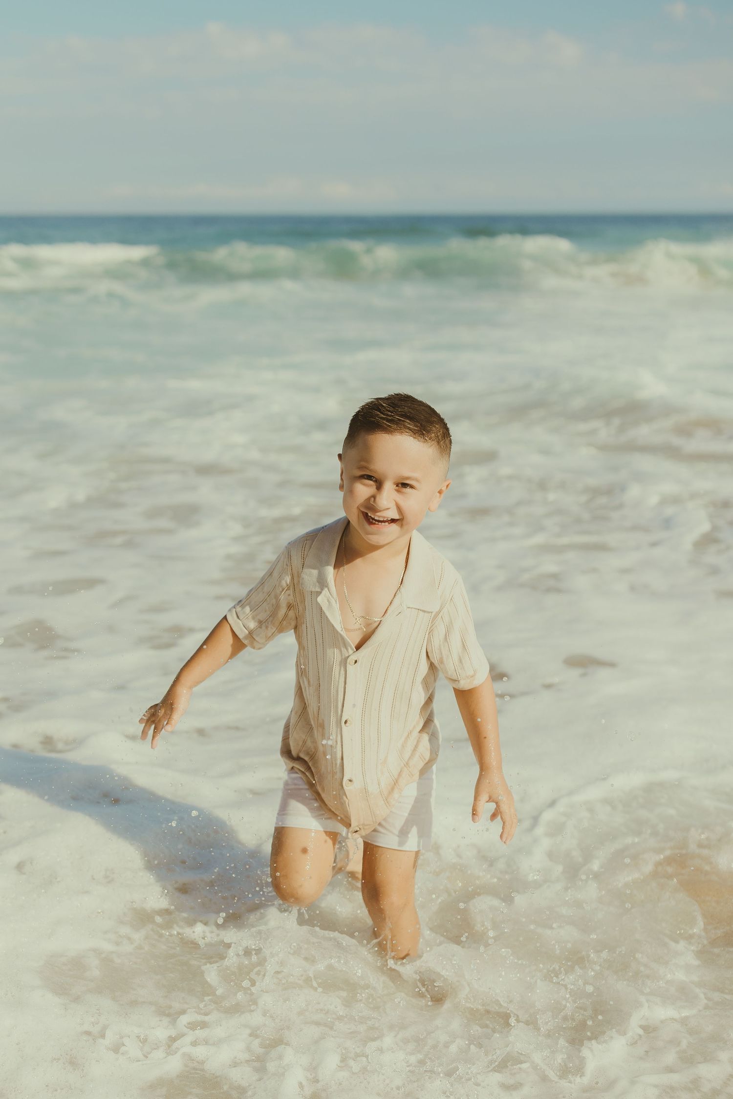 A young child plays in ocean waves at the beach wearing a beige button-up shirt and white shorts.