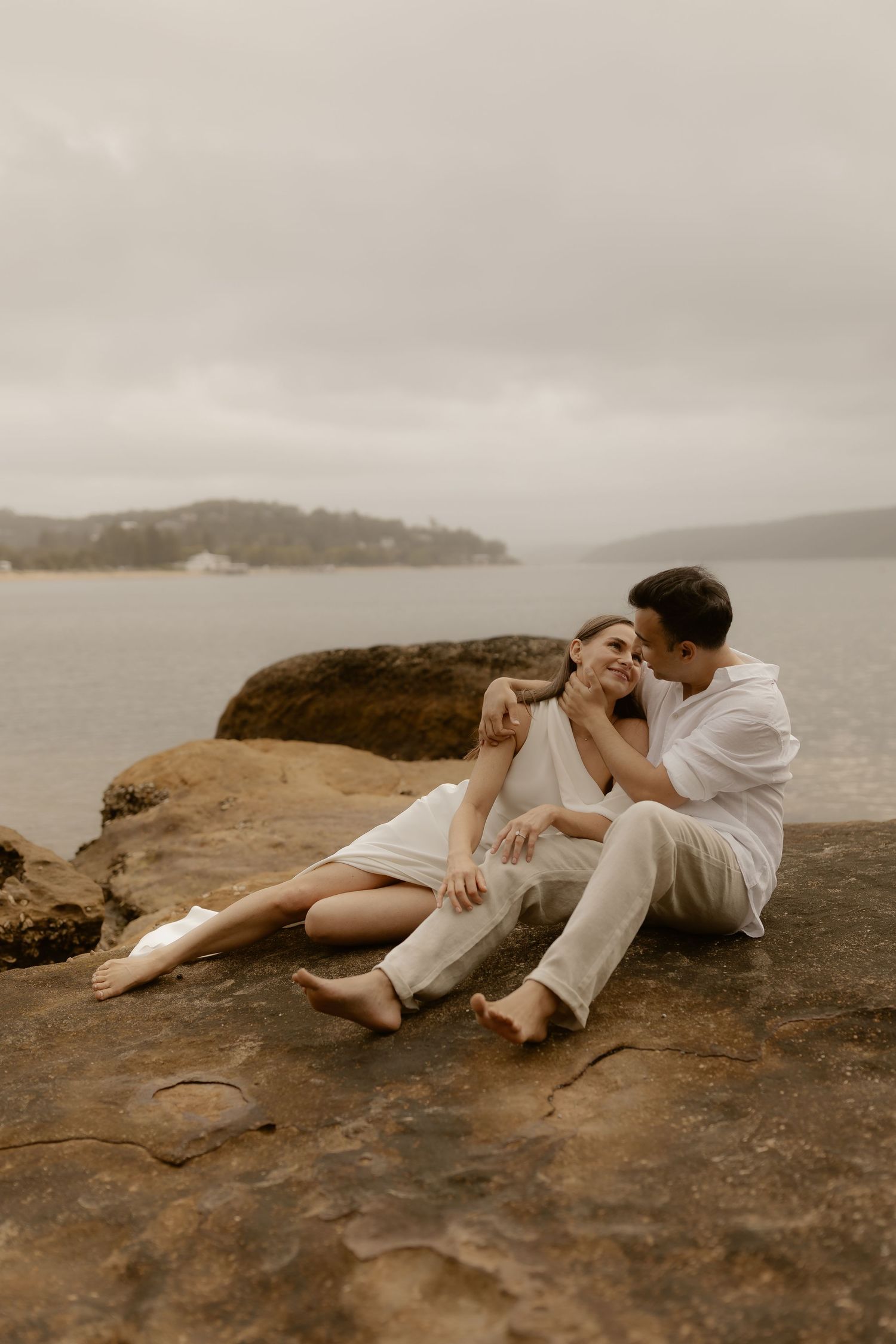 Romantic couple in white clothing lying together on coastal rocks with ocean view in moody lighting.