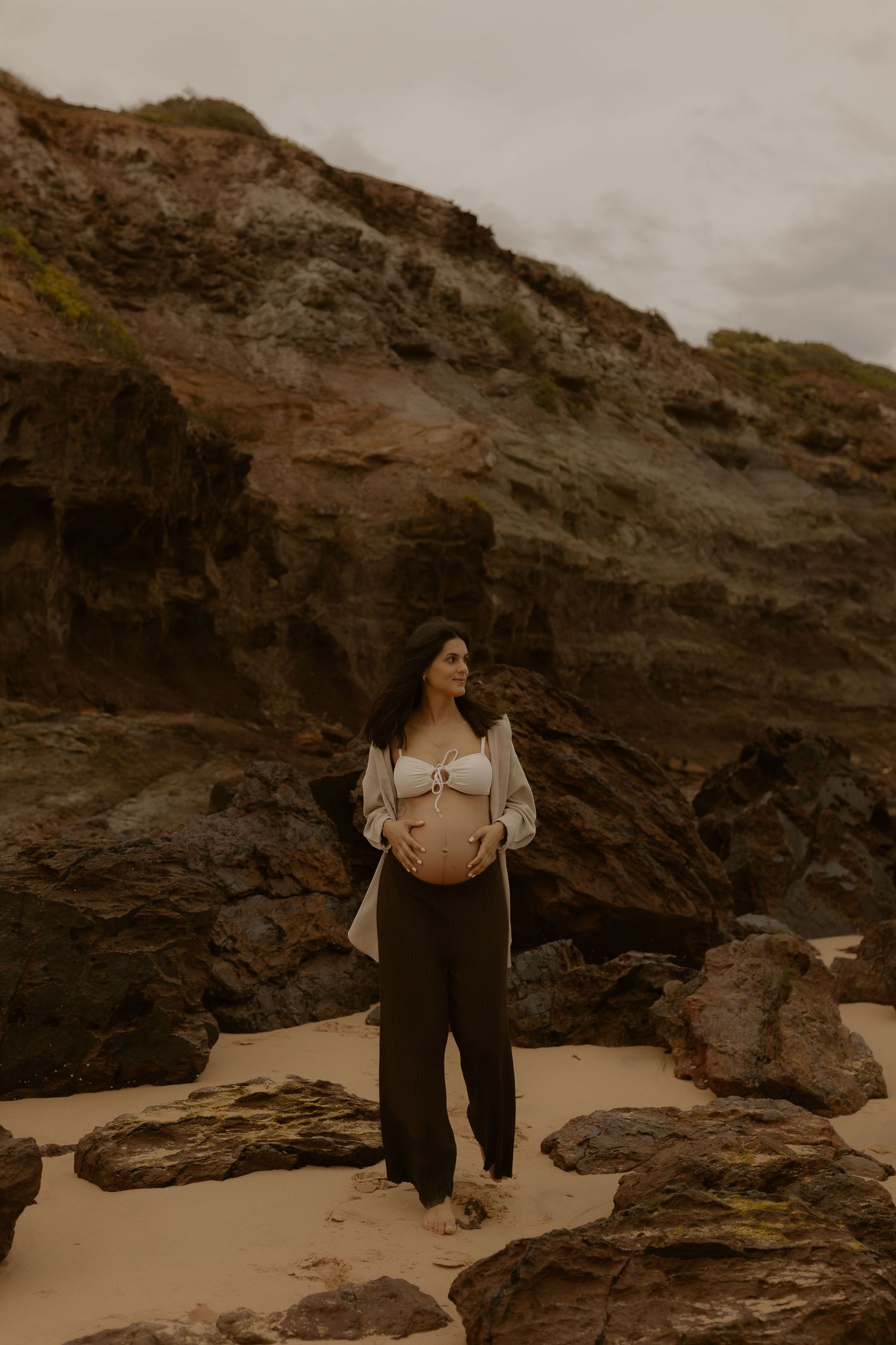 A moody beach maternity photoshoot with dramatic rocky cliffs and overcast skies in the background.