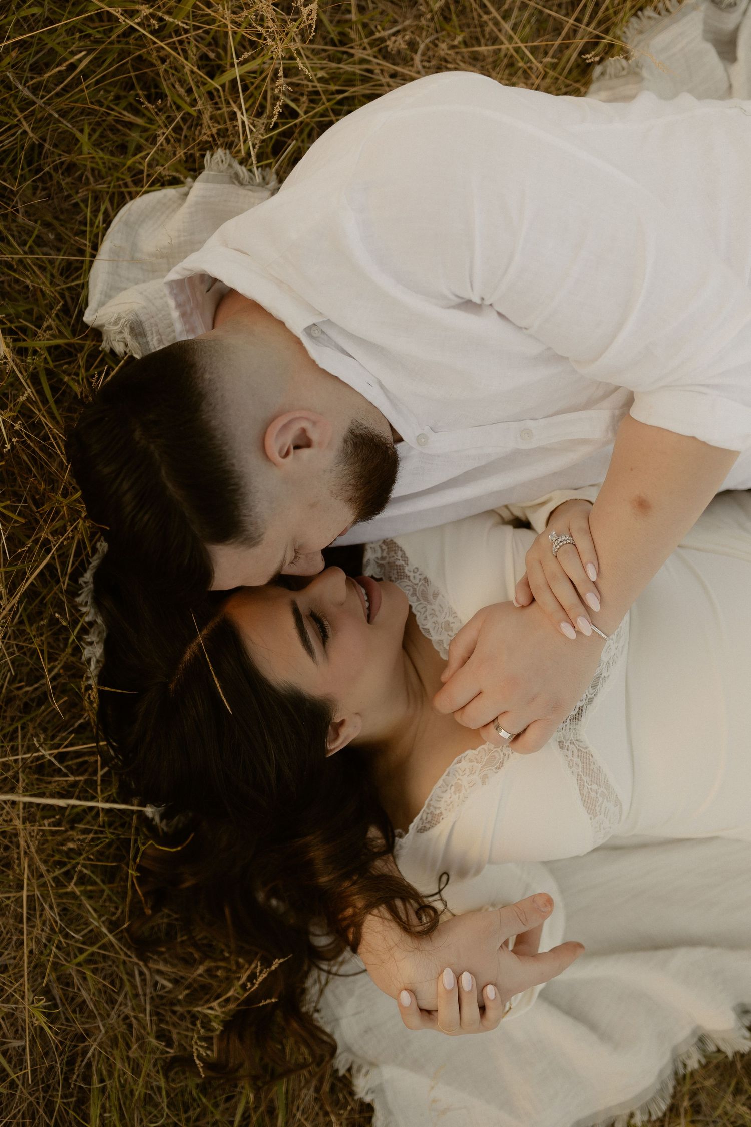A couple in white clothing lies together intimately in a field of tall grass during golden hour.