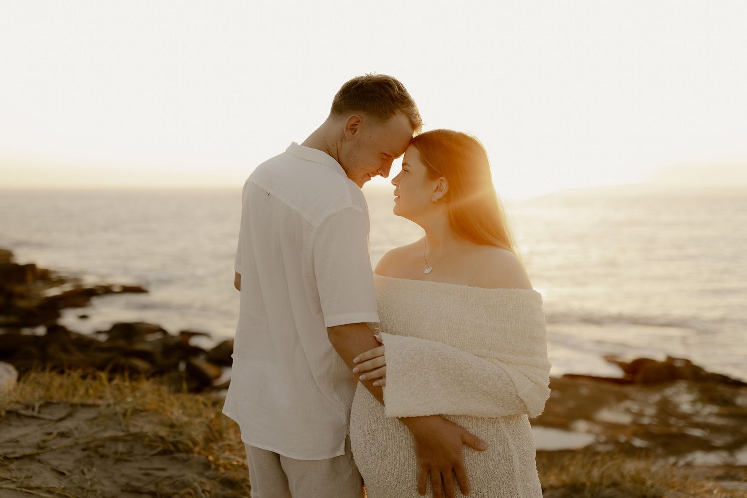 Couple in white outfits sharing romantic moments during sunset at a scenic coastal location.
