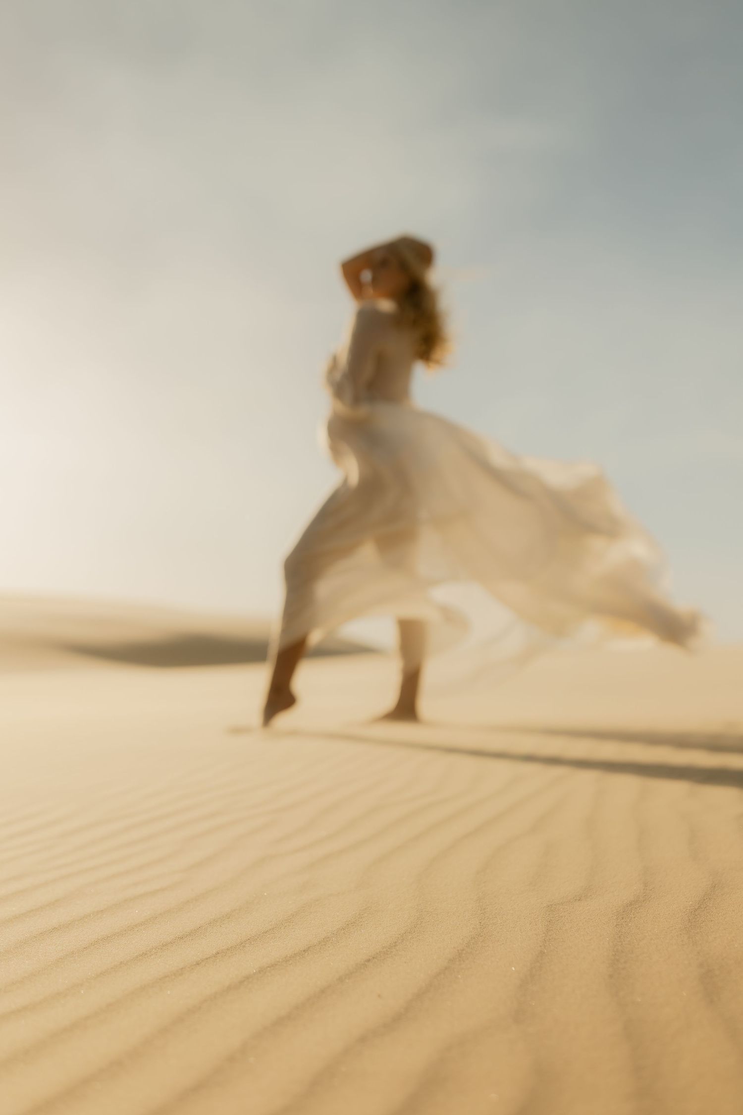 A woman in a flowing white dress runs across golden sand dunes under a hazy sky in a dreamy, blurred desert scene.